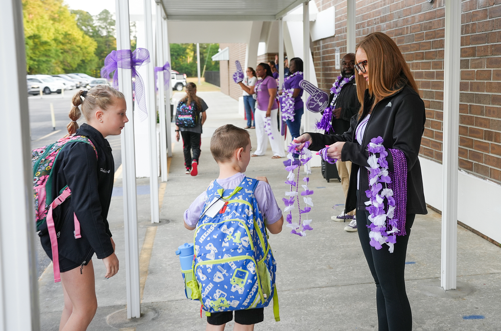 Adults and children wearing purple leis and carrying backpacks walk past each other on a sidewalk.