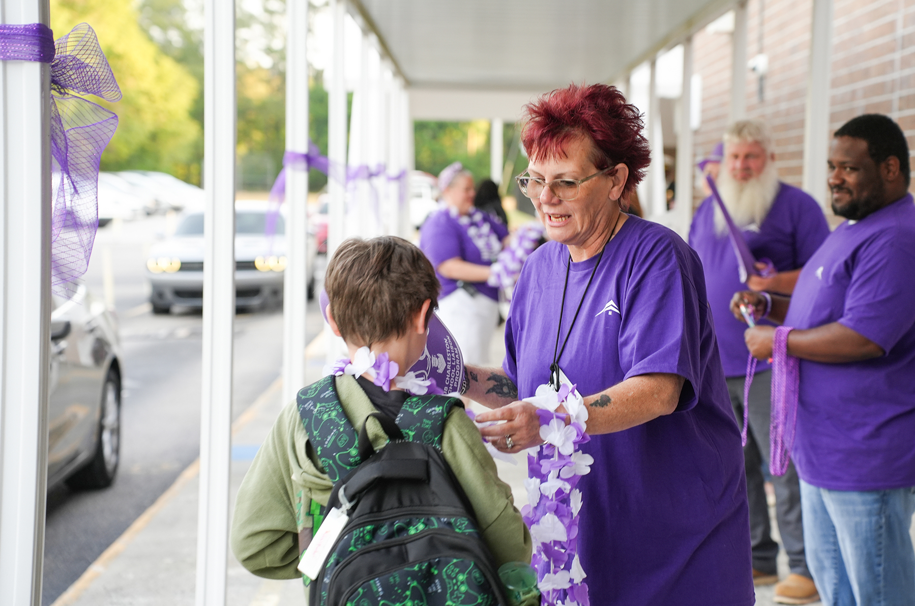 A group of people in purple shirts on a porch. A woman hands a child a lei.