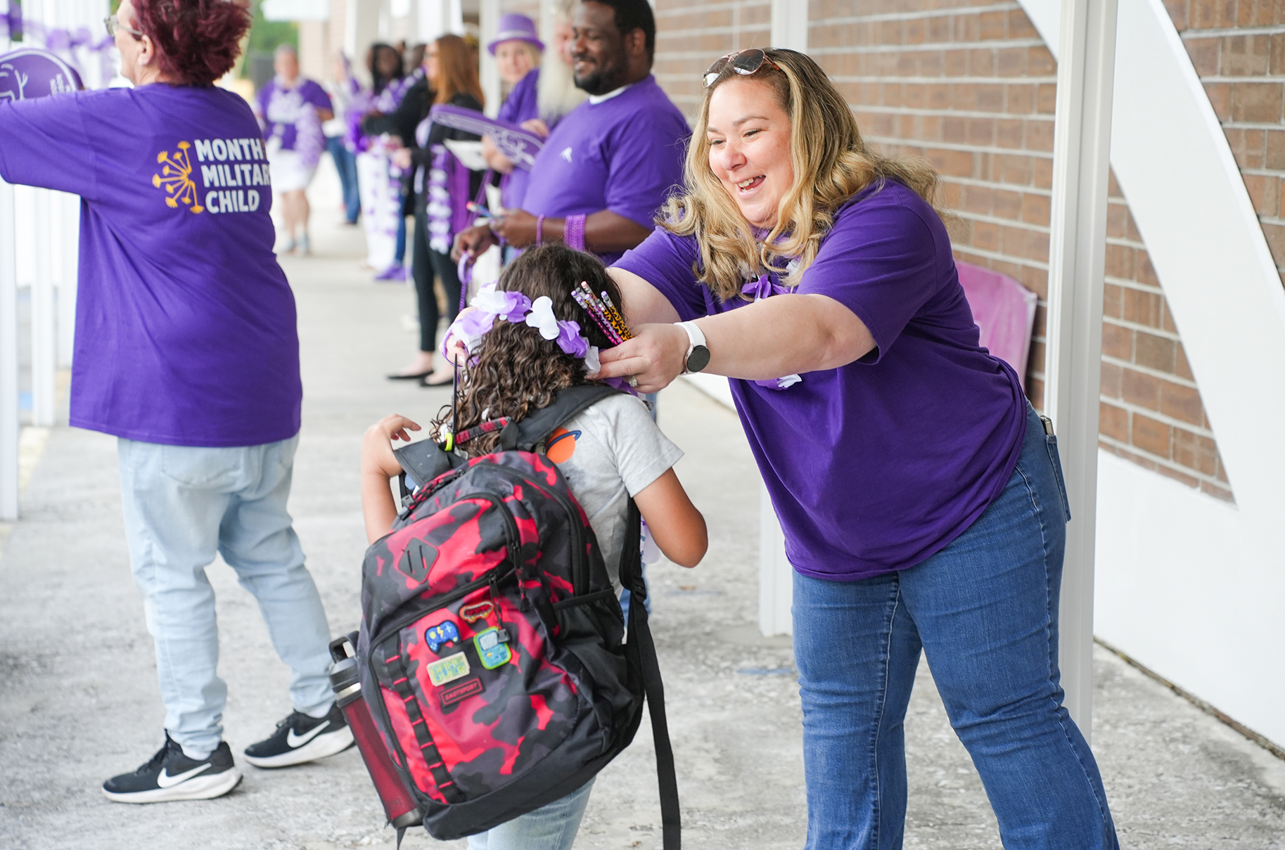 A woman helps a child with a backpack, both wearing purple shirts. A person stands in the background with a purple shirt and a crowd in the distance.