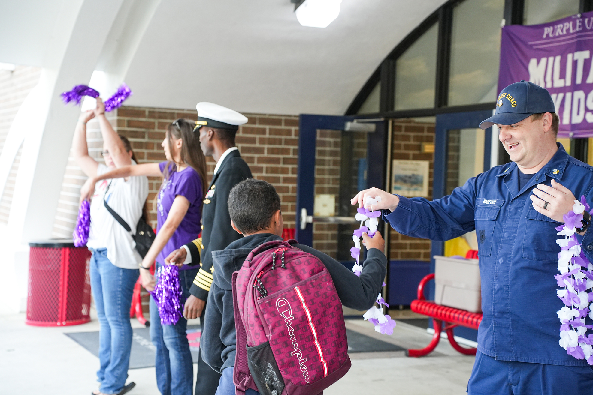A uniformed man hands a lei to a boy wearing a backpack. Several people stand around them.