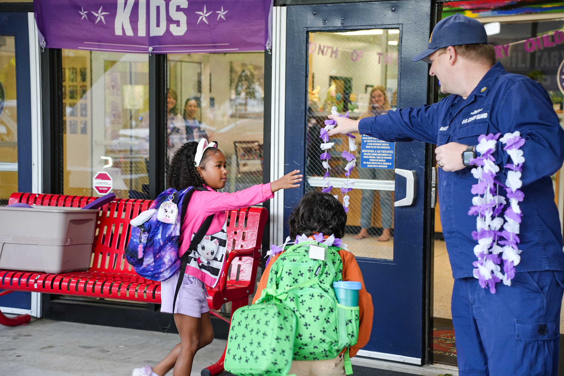 Two children, one with a backpack, receiving leis from a uniformed person by a red bench.