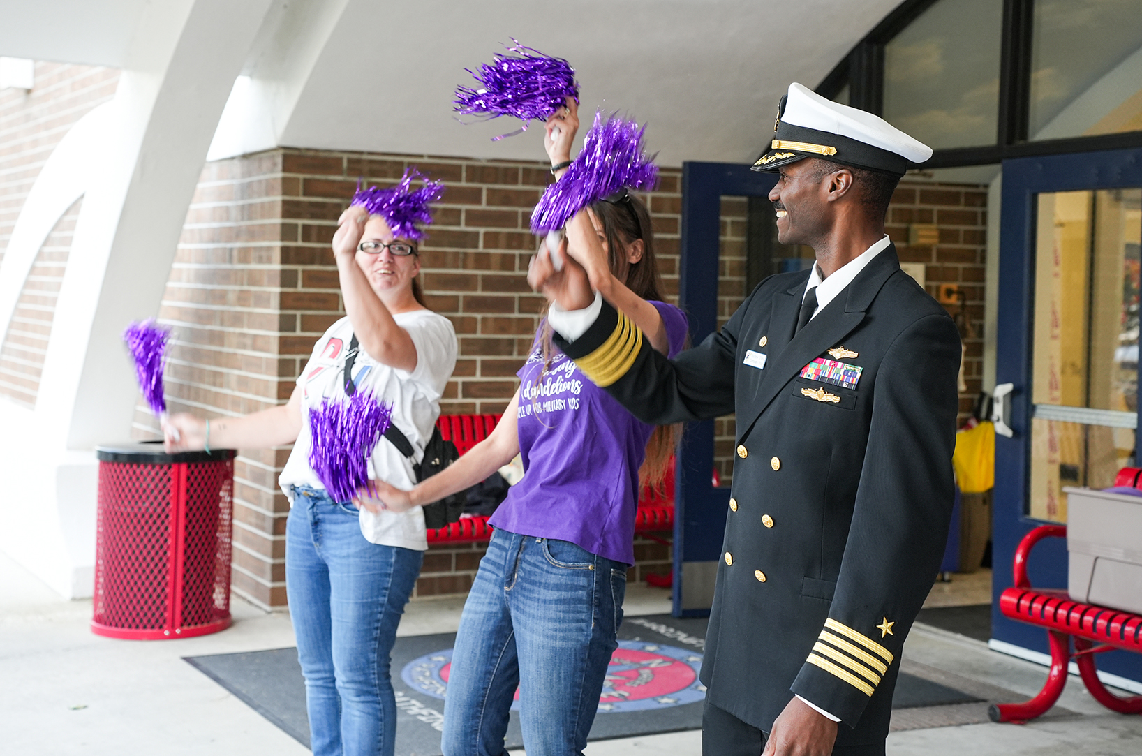 Two women holding purple pom-poms stand near a uniformed man in a military hat. They are outside a building with a red bench in the background.