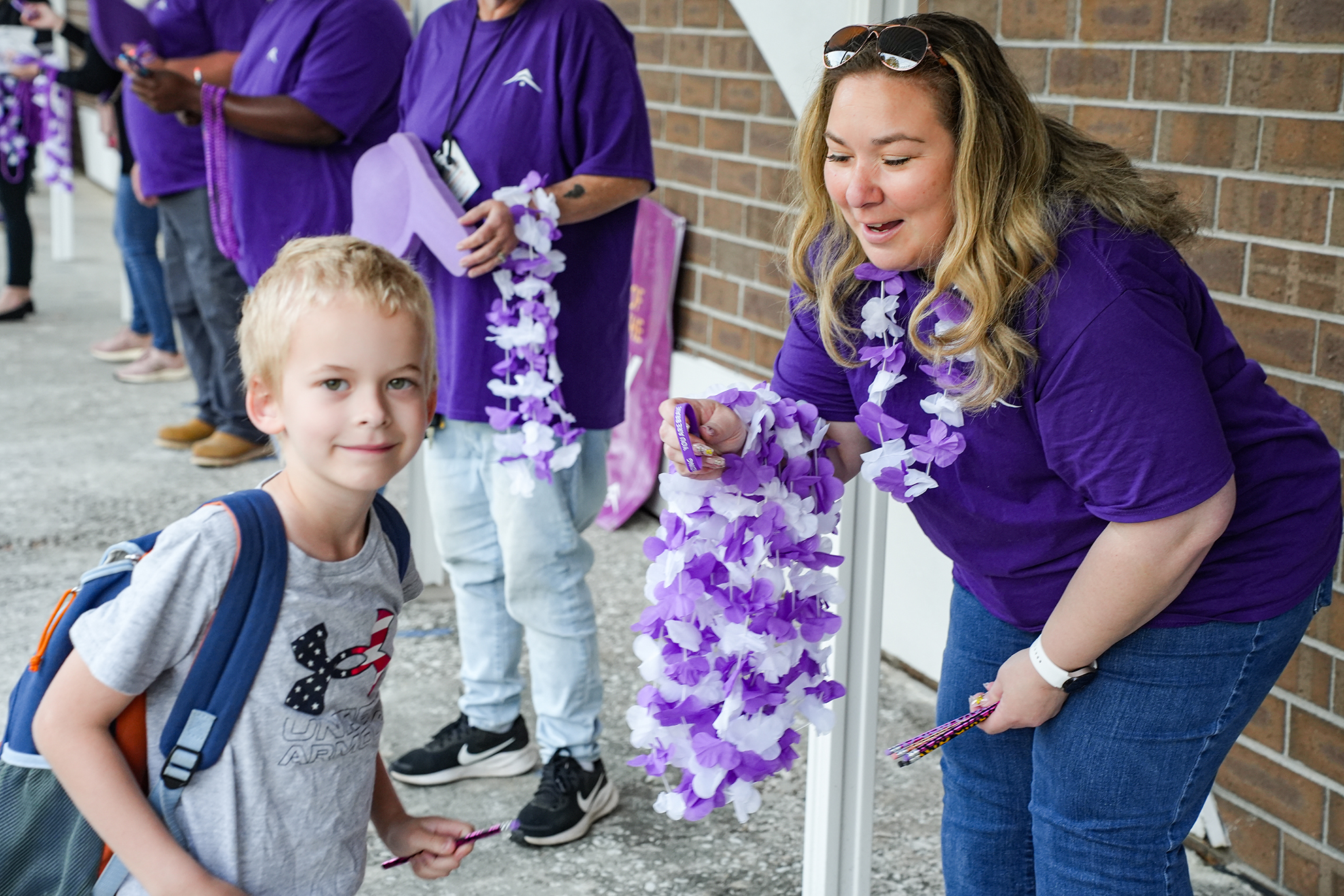 A woman in a purple shirt places a lei on a child wearing a gray shirt. Behind them are other adults wearing purple.