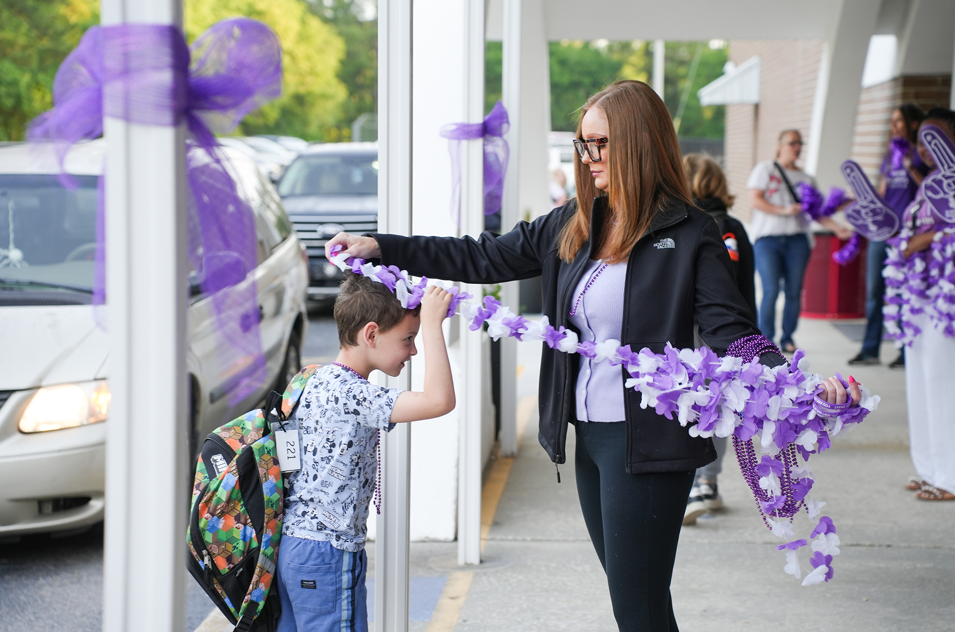 A woman helps a child hold a purple ribbon while a car is parked in the background.