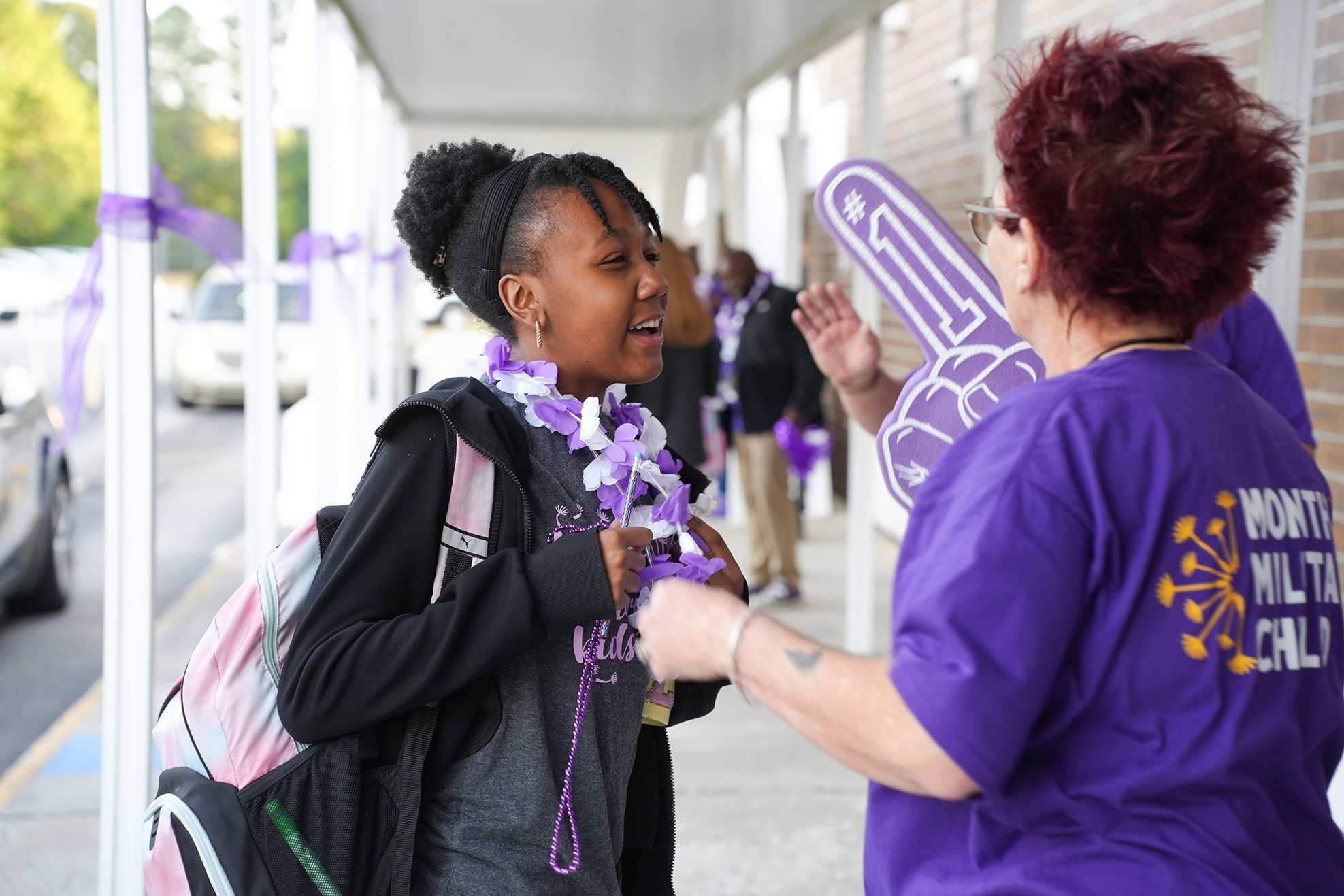 A woman talks with a young girl wearing a purple ribbon, with a building and parked cars in the background.