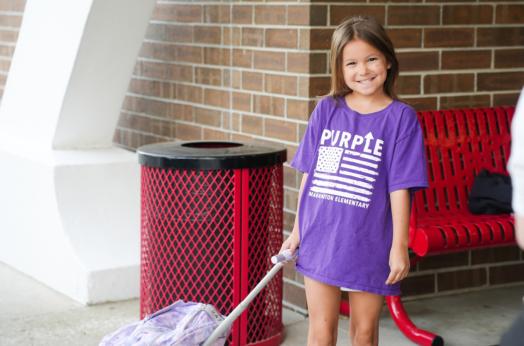 A young girl in a purple T-shirt stands by a red trash can and bench. She is pulling a small bag.