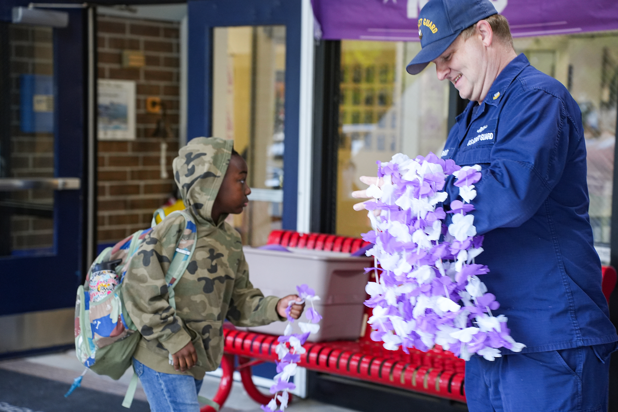 Person in navy uniform hands purple flowers to a child with a backpack in front of a red bench.