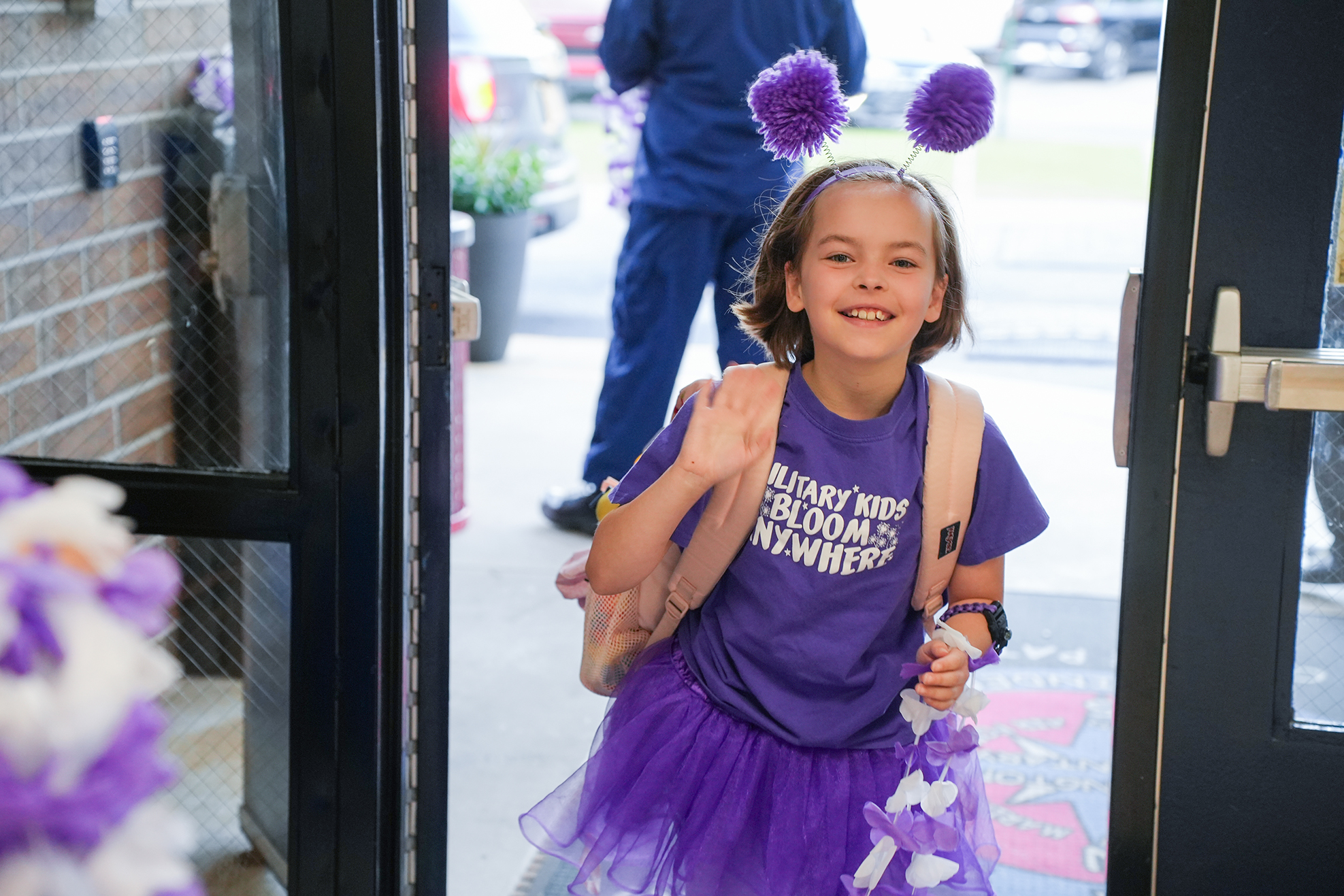 A young girl in a purple tutu with purple pom-poms on her head, smiling, stands in an open doorway.