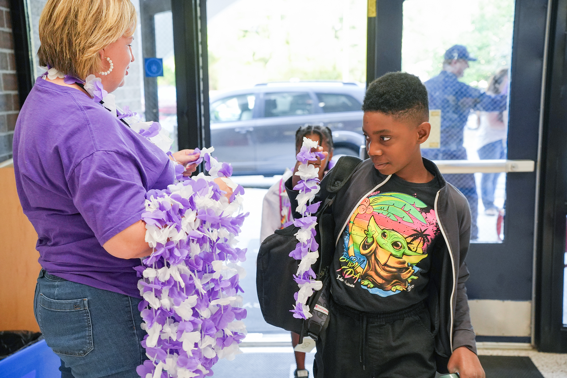 Woman in purple gives a young boy a lei. They stand in a doorway with sunlight.