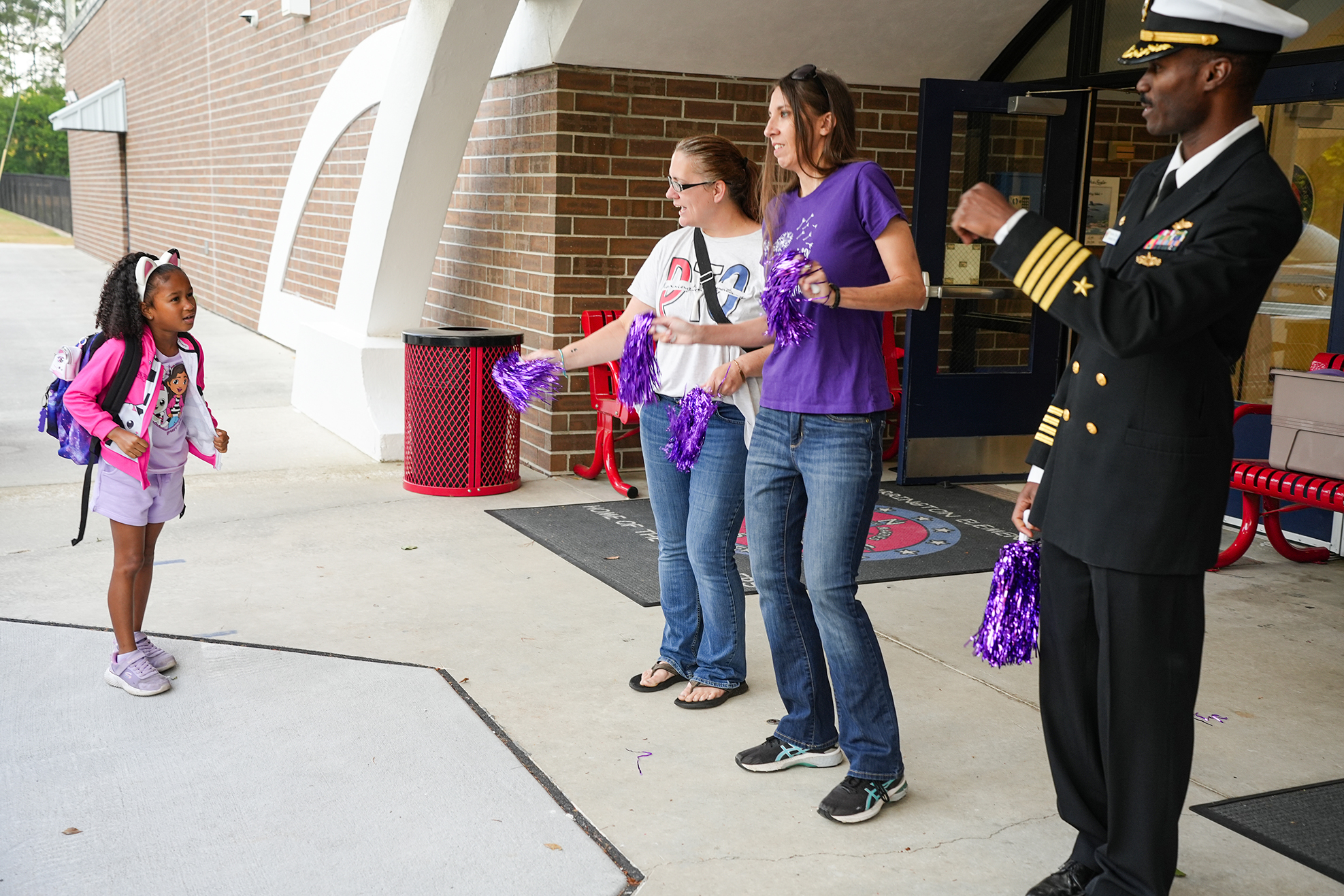 Four people stand outside a building, one in uniform, while a young girl in front holds purple ribbons.