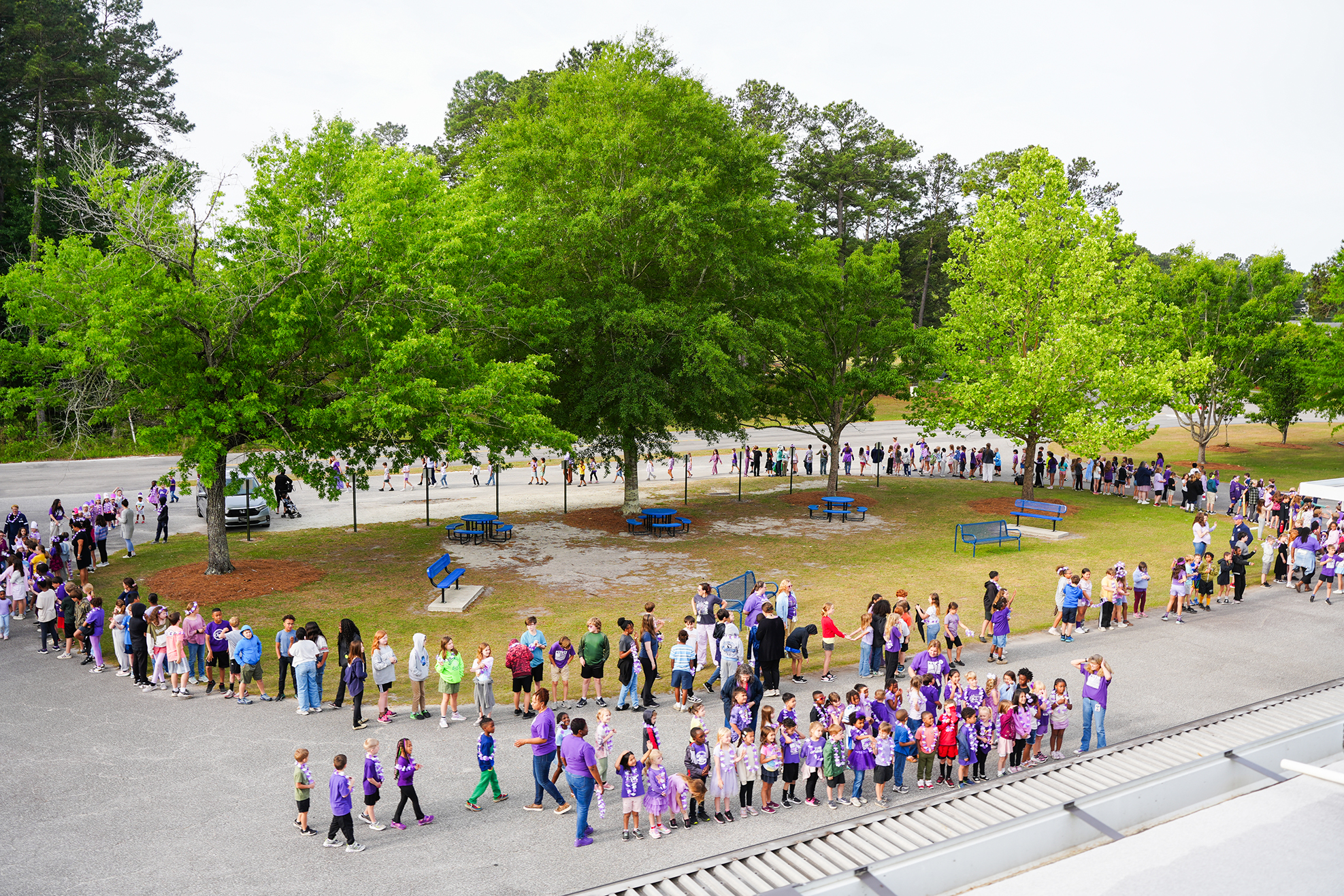 A group of people, some wearing purple, standing in a circle in a park with green trees and picnic tables.