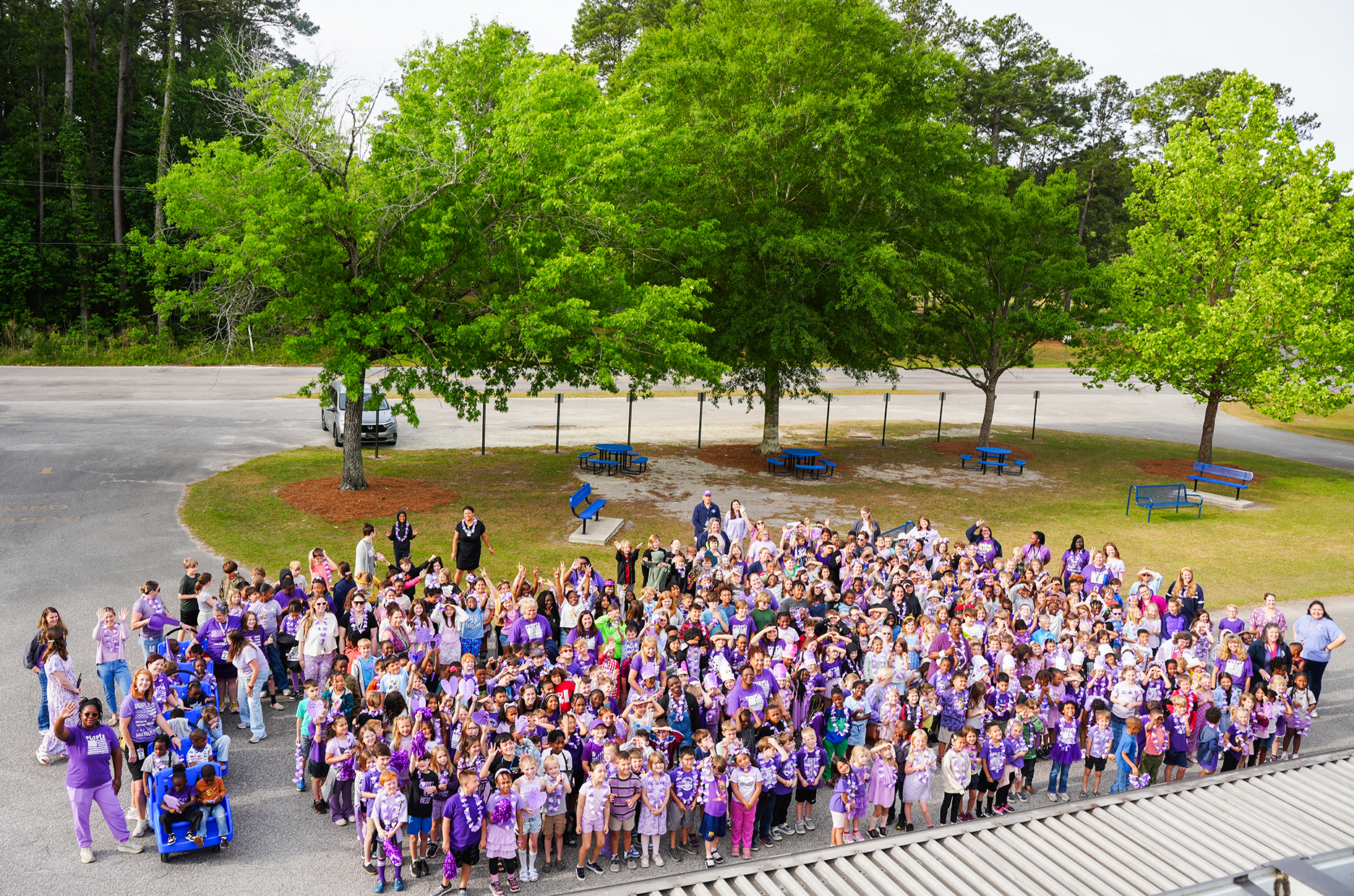 A large group of people wearing purple shirts gathers in a parking lot with trees and benches.