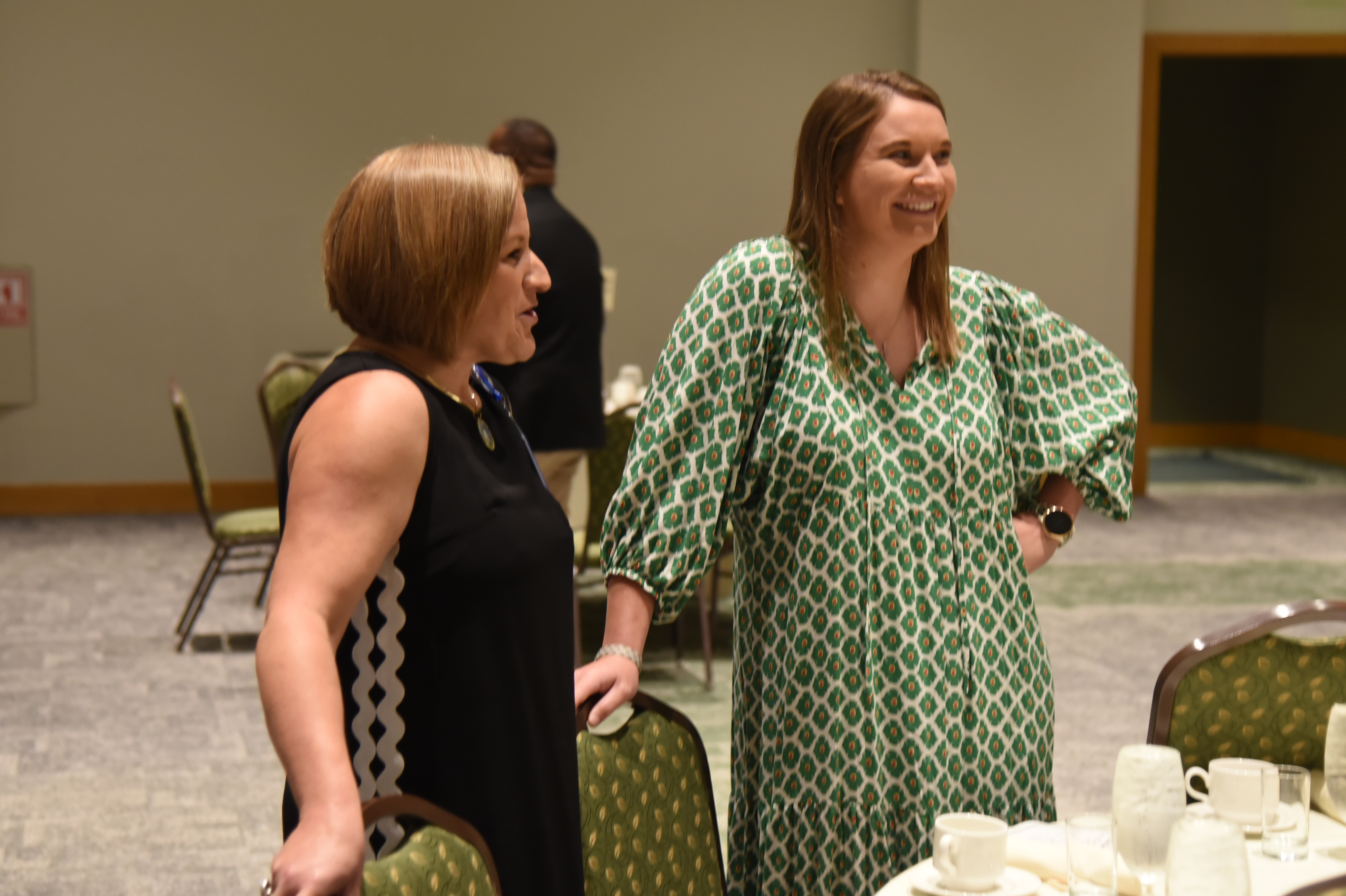 Two women converse in a room with tables and chairs. One wears a black dress, the other a green dress.