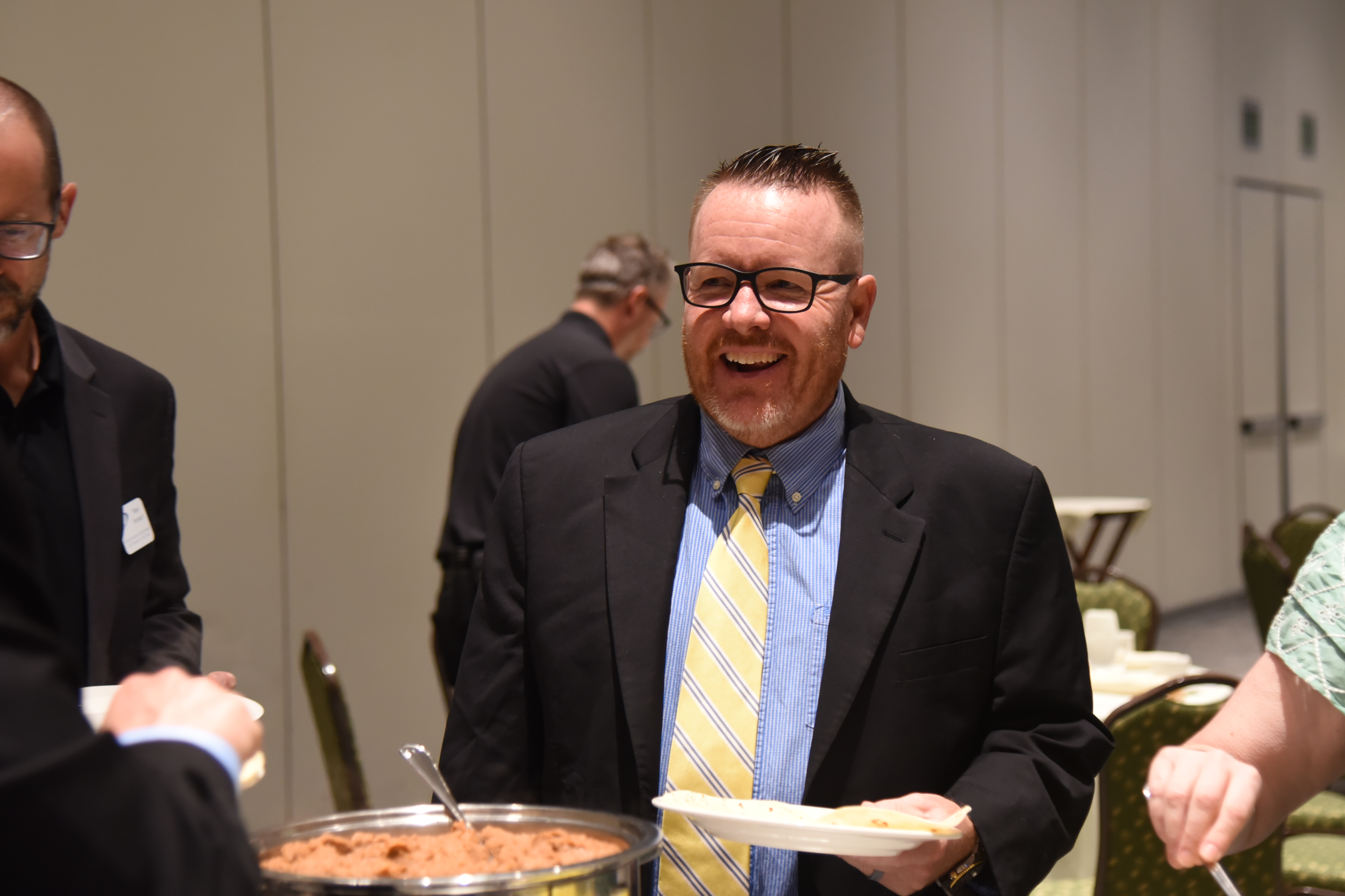 A man in a suit and tie stands at a buffet table, holding a plate of food.
