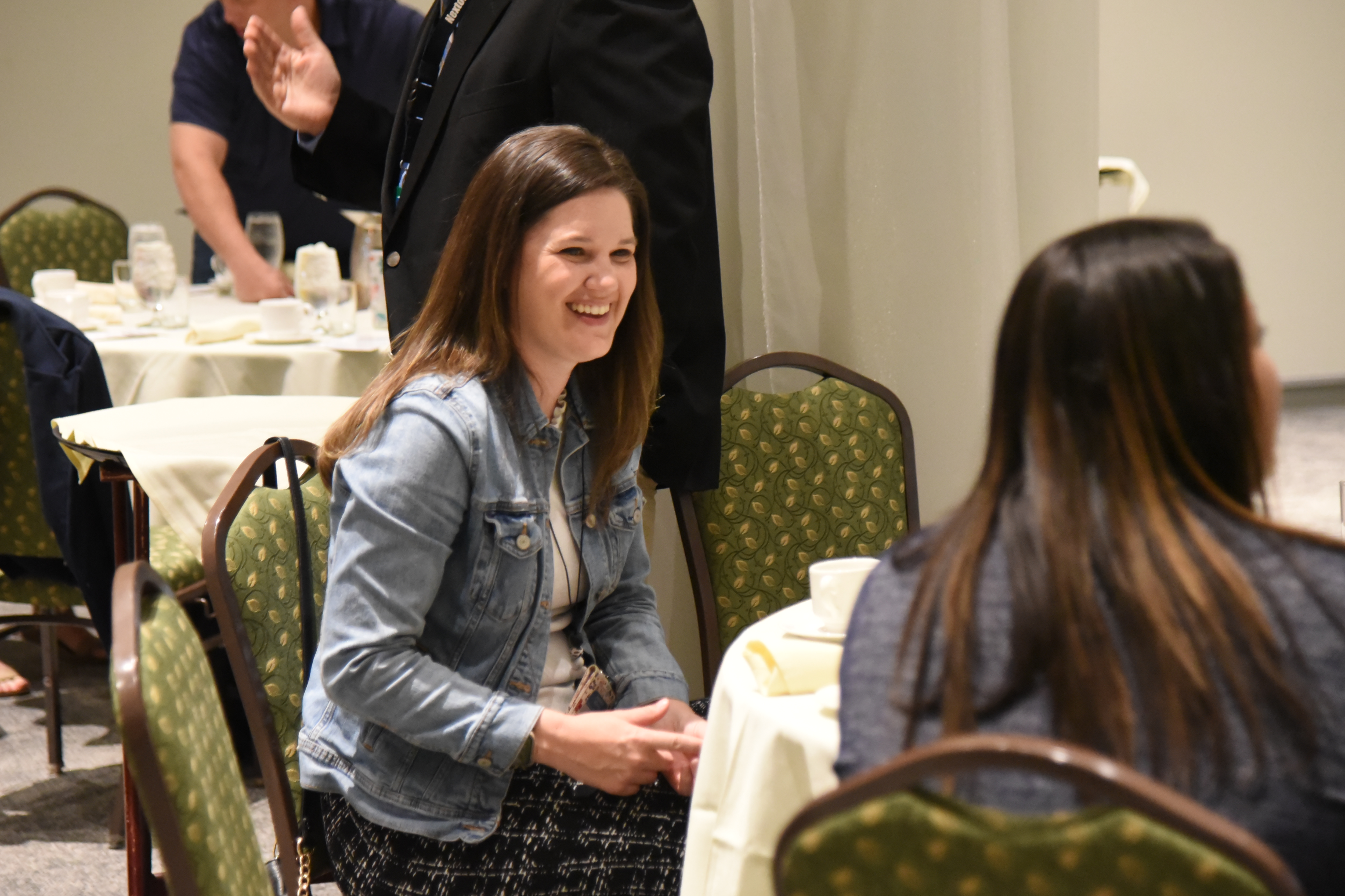 A woman in a denim jacket smiles at another person in a restaurant. They sit at a table with white cloth.