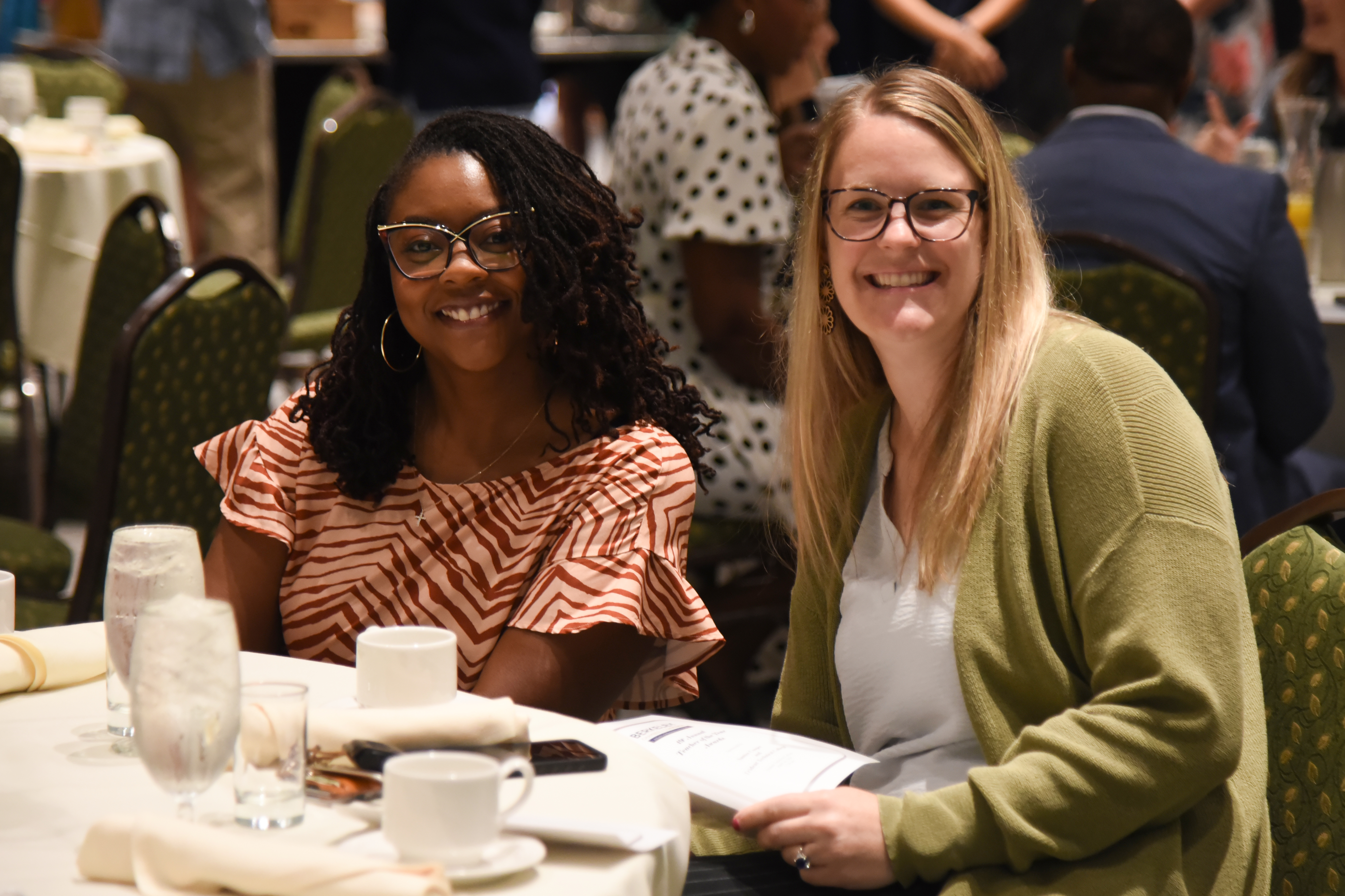 Two women in glasses sit at a table. One wears a white shirt and green cardigan, the other a red shirt.
