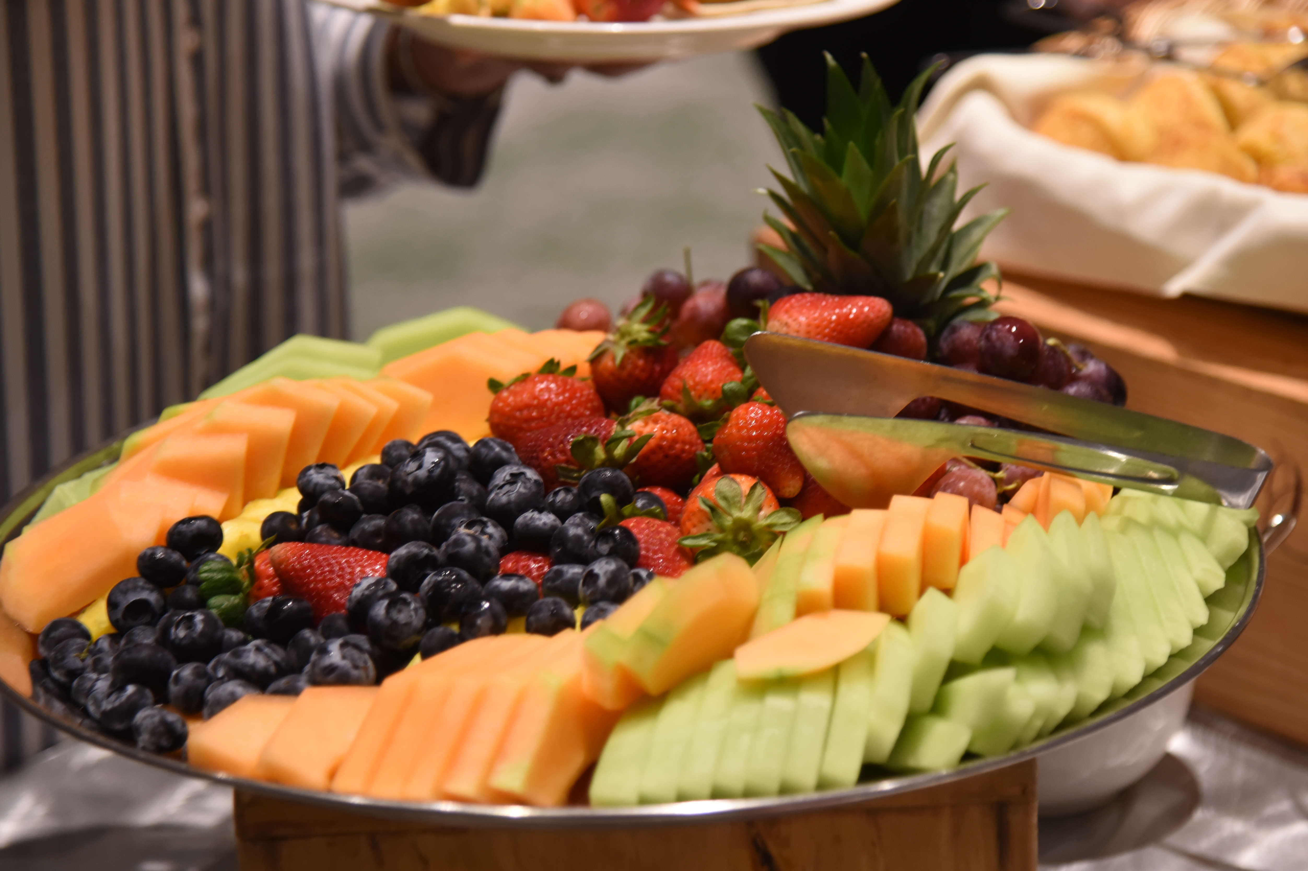 A large round tray displays various fruits, including strawberries, blueberries, cantaloupe, pineapple, and grapes.
