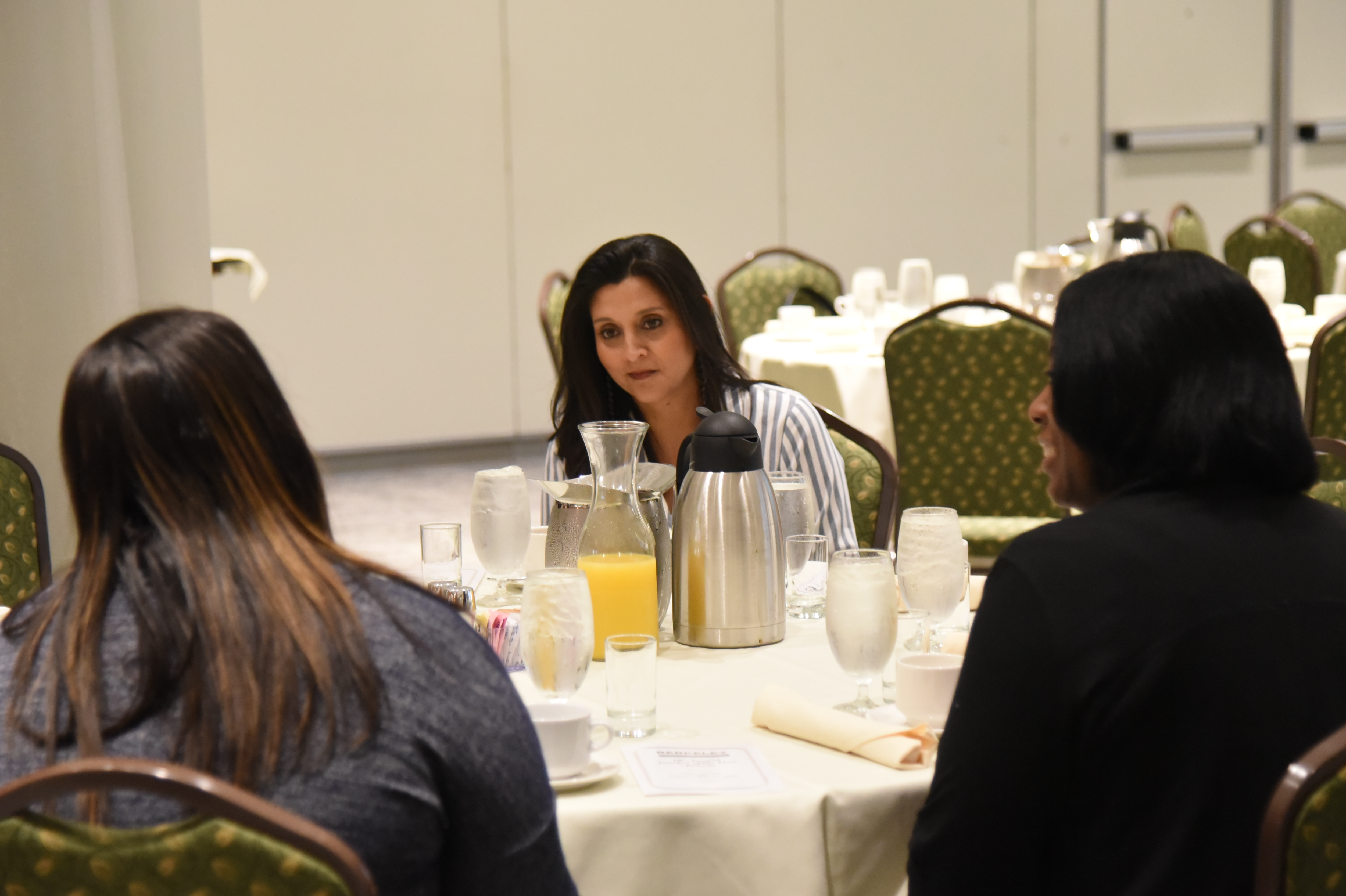 Three people sit at a table, facing each other. A woman in a striped shirt speaks. Glassware, a silver pitcher, and a napkin are on the table.