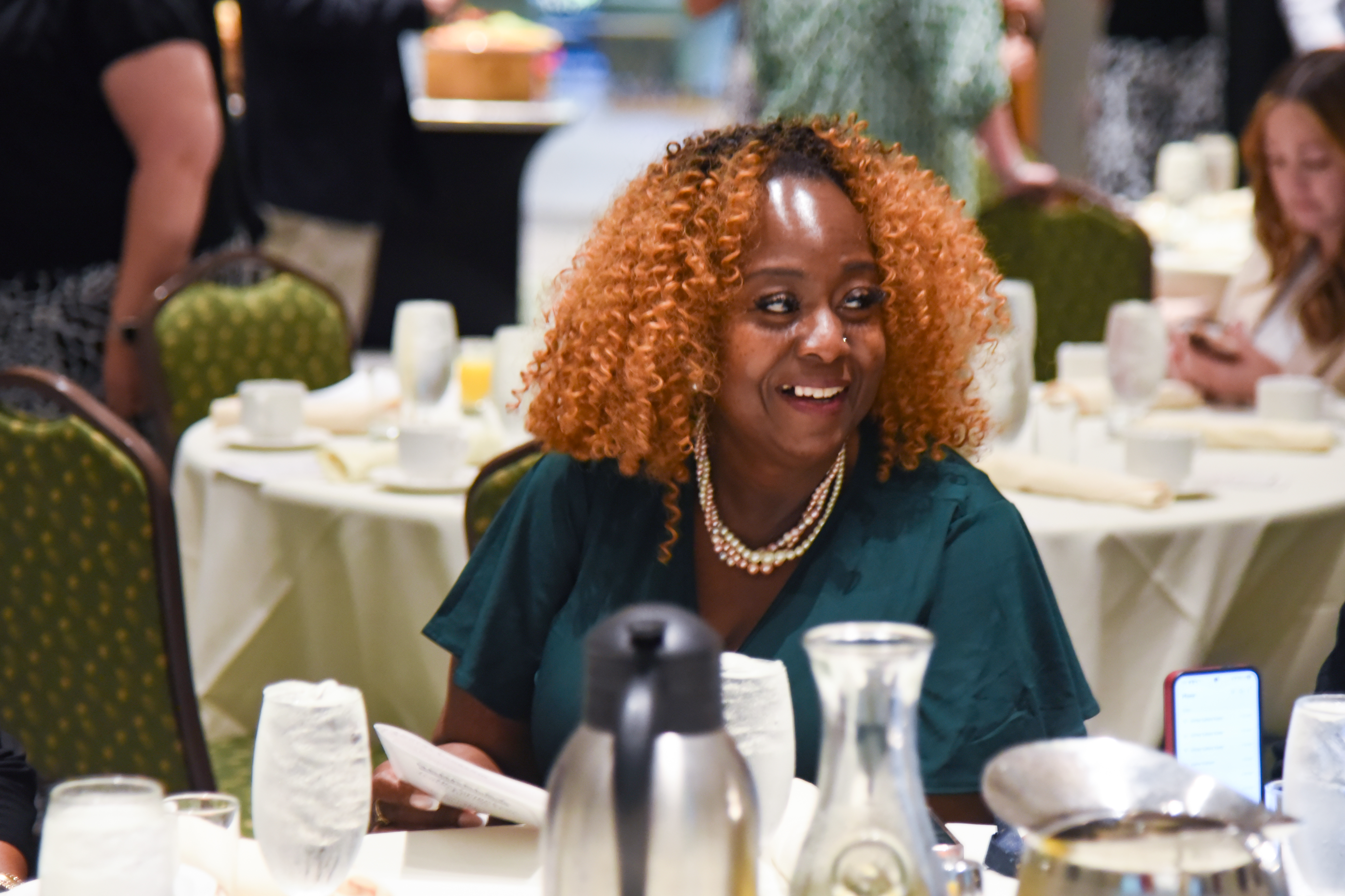A person with curly hair, wearing a necklace, sits at a table with a coffee pot.