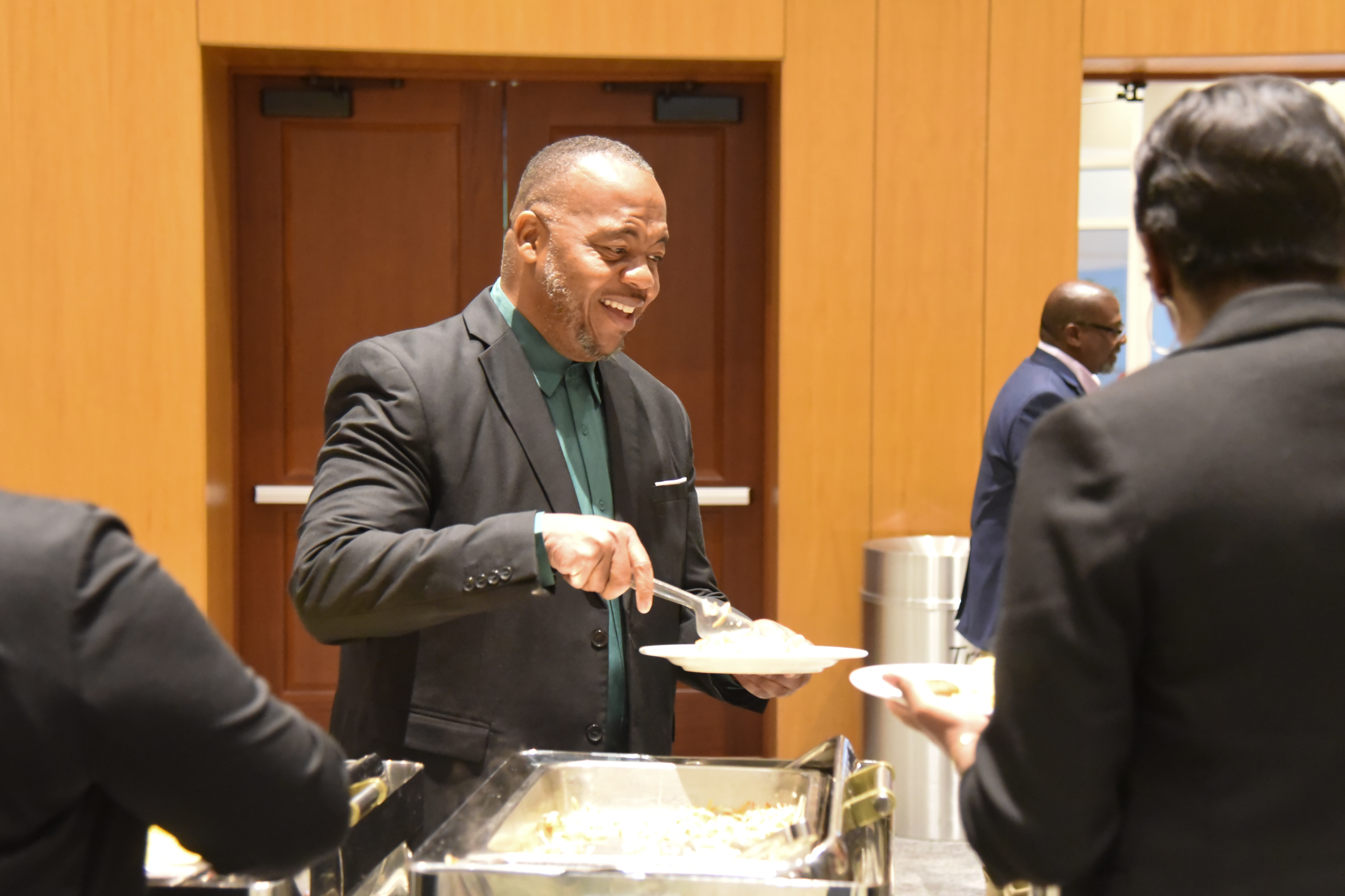 Man in a green shirt serving food to two people standing at a buffet table.