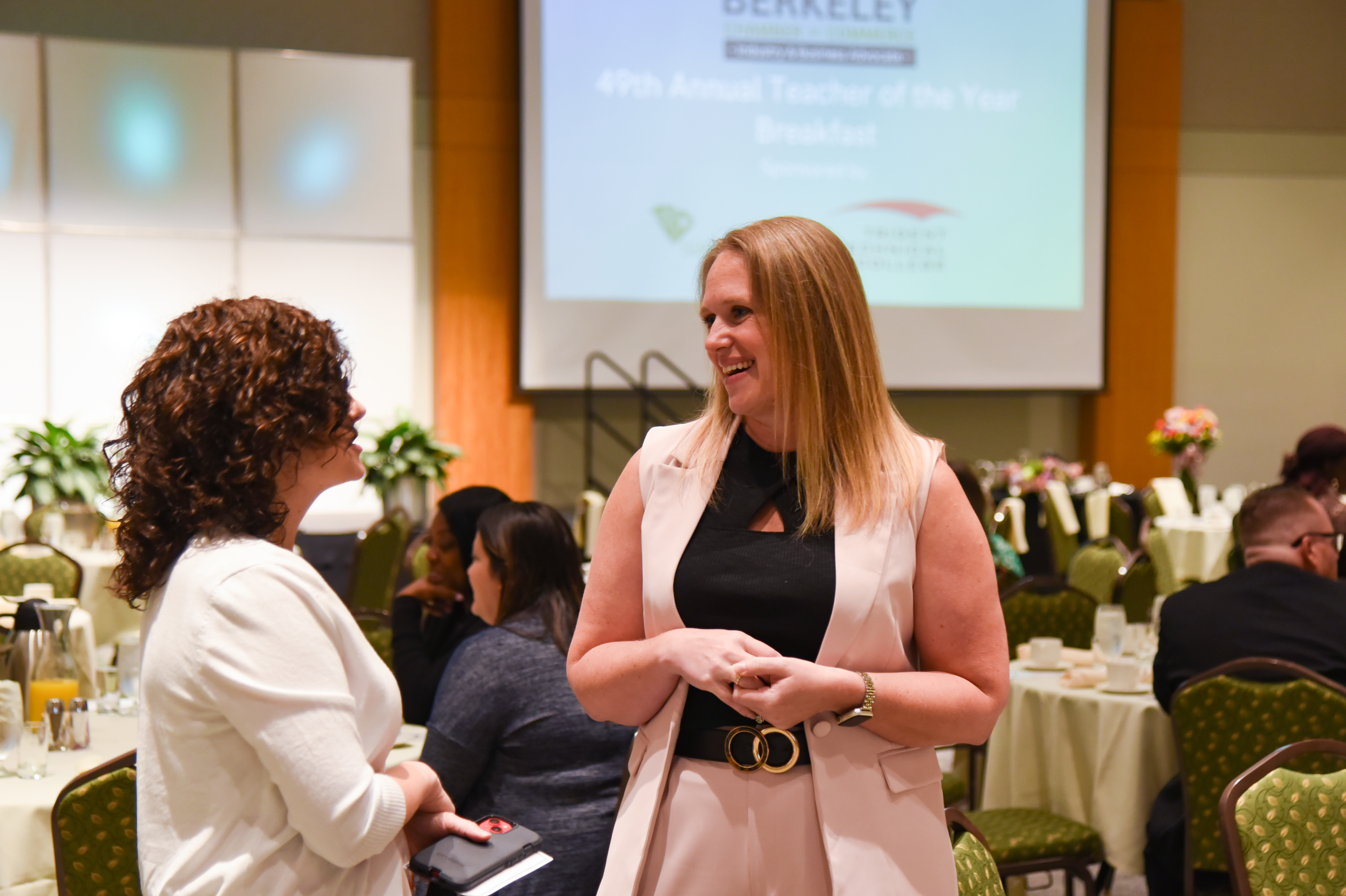 Two women in formal attire converse near a presentation screen reading "40th Annual Teacher of the Year Breakfast."