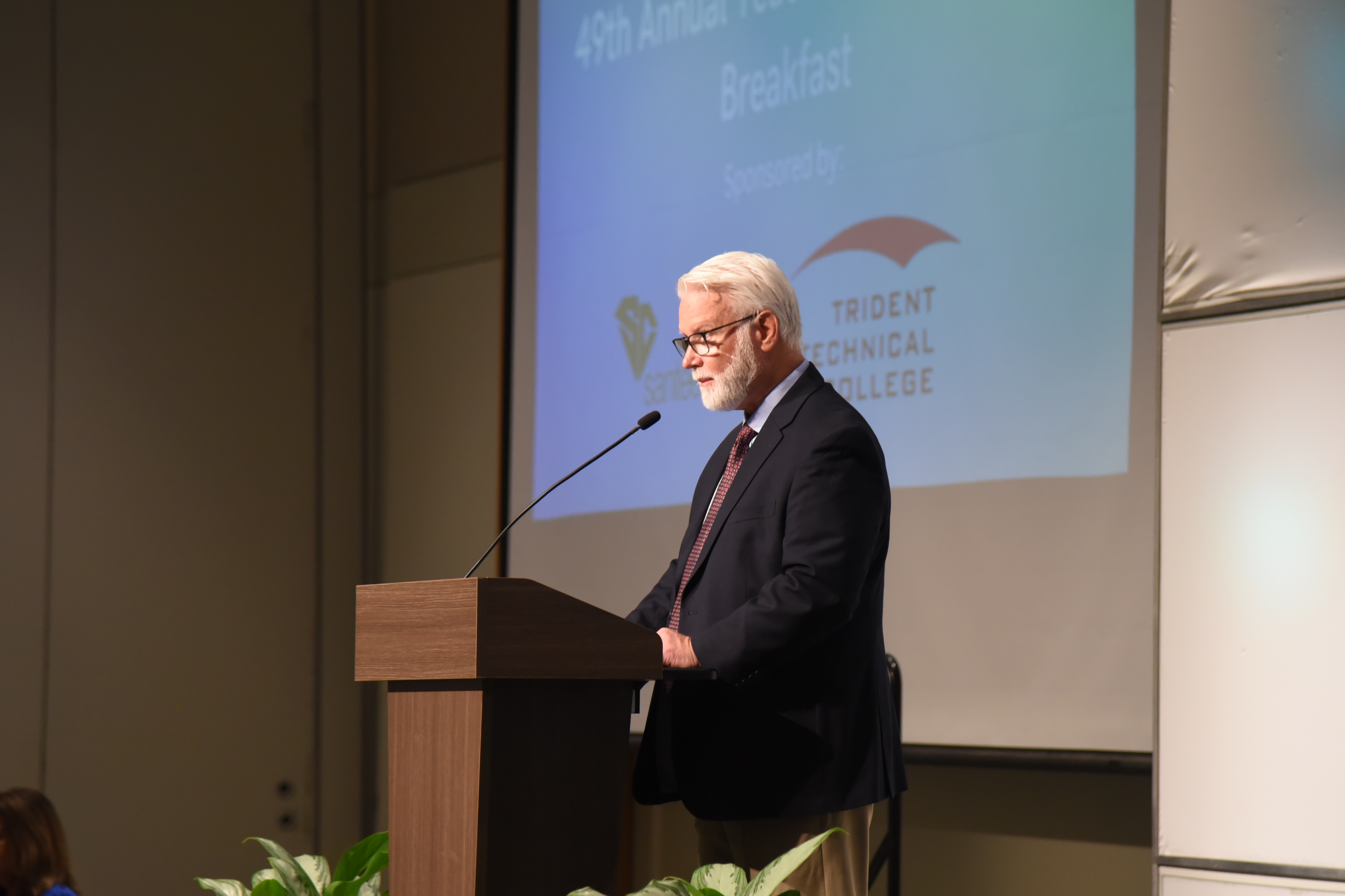 A man in a suit stands at a podium with a microphone in a room with a large screen behind him.