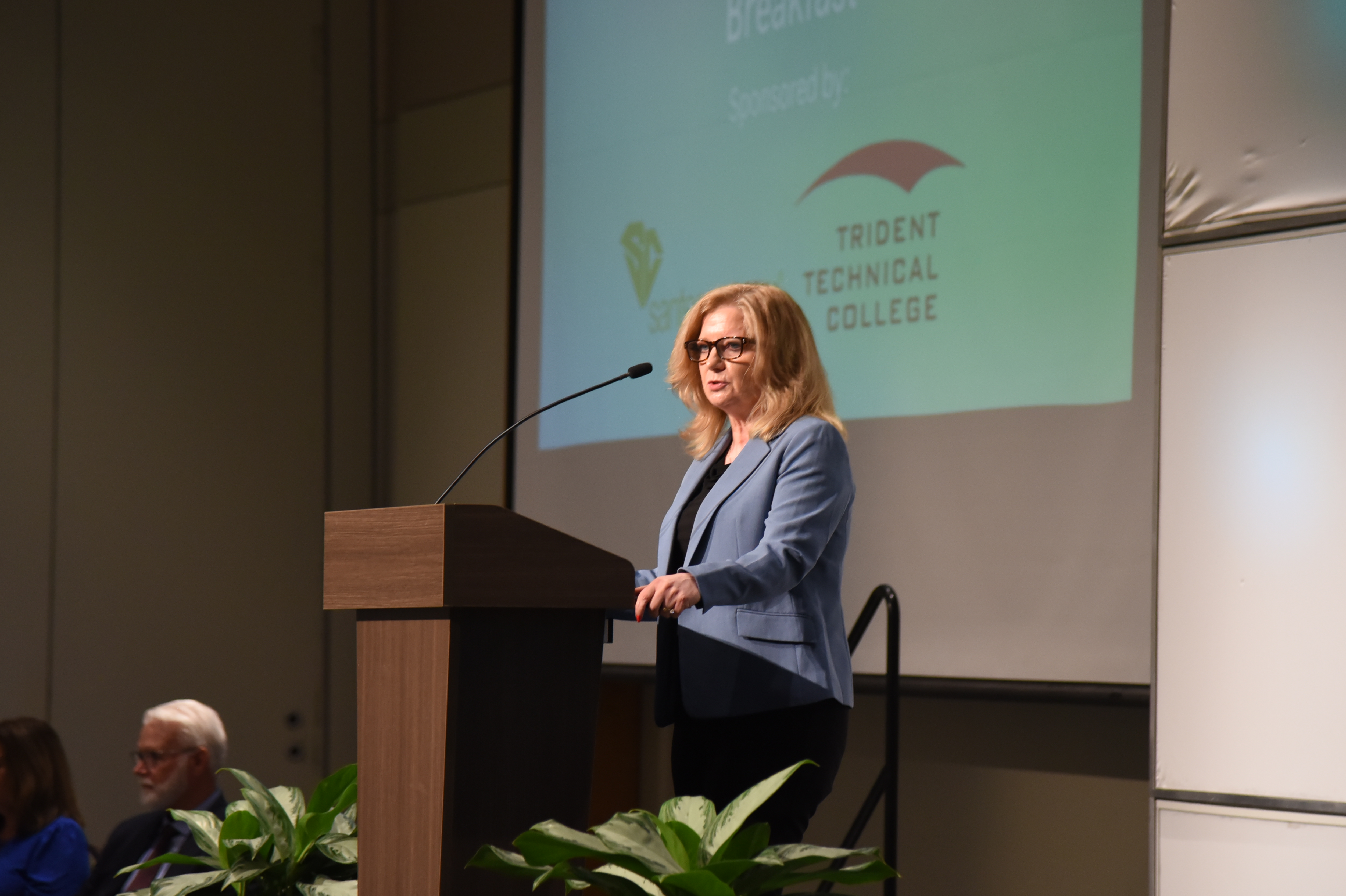 Woman stands at podium with microphone in front of a projector screen. Behind her, people sit in chairs.