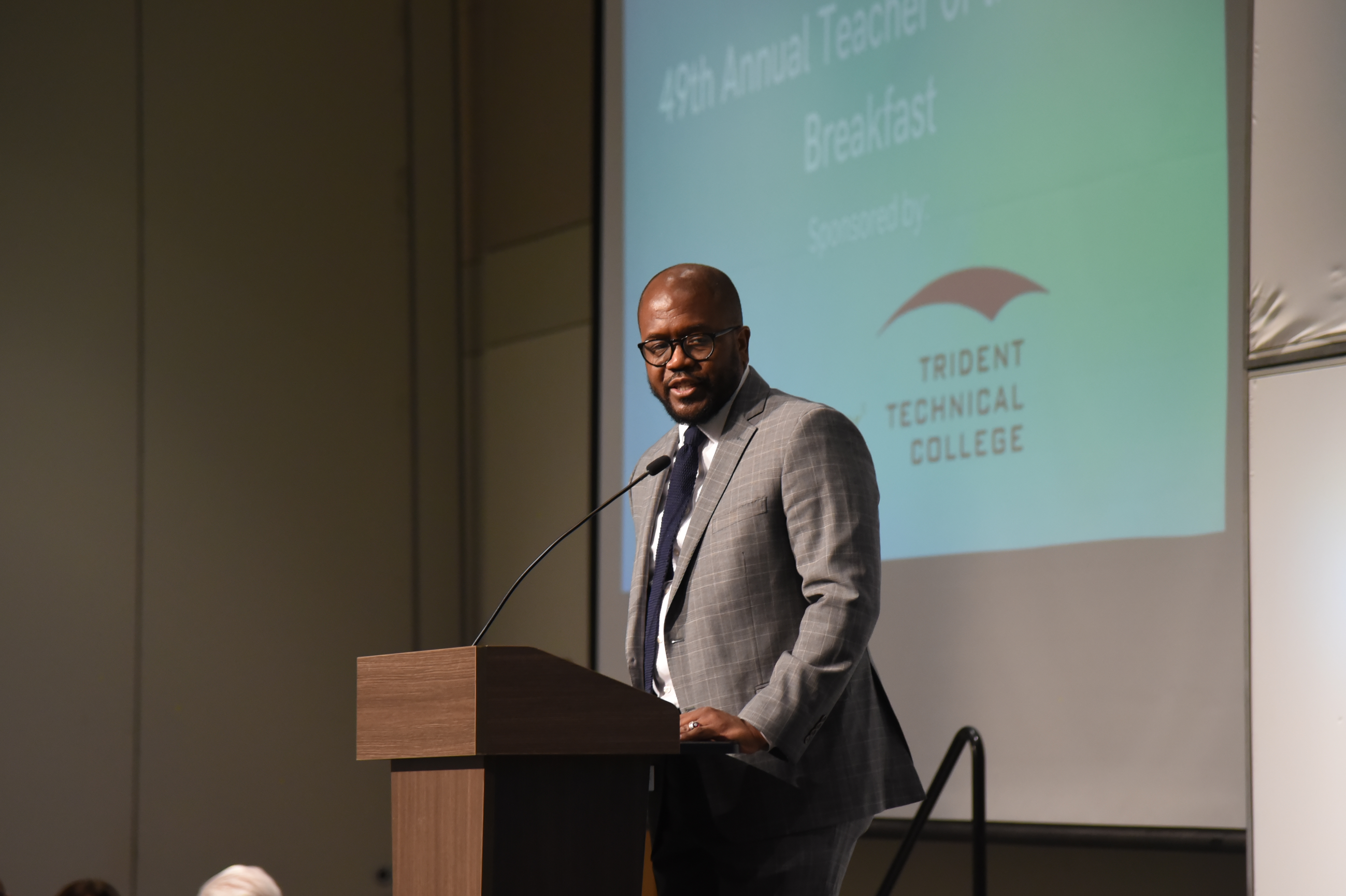 A person stands at a podium with a microphone. Behind, a projector screen reads "49th Annual Teacher of the Year Breakfast".