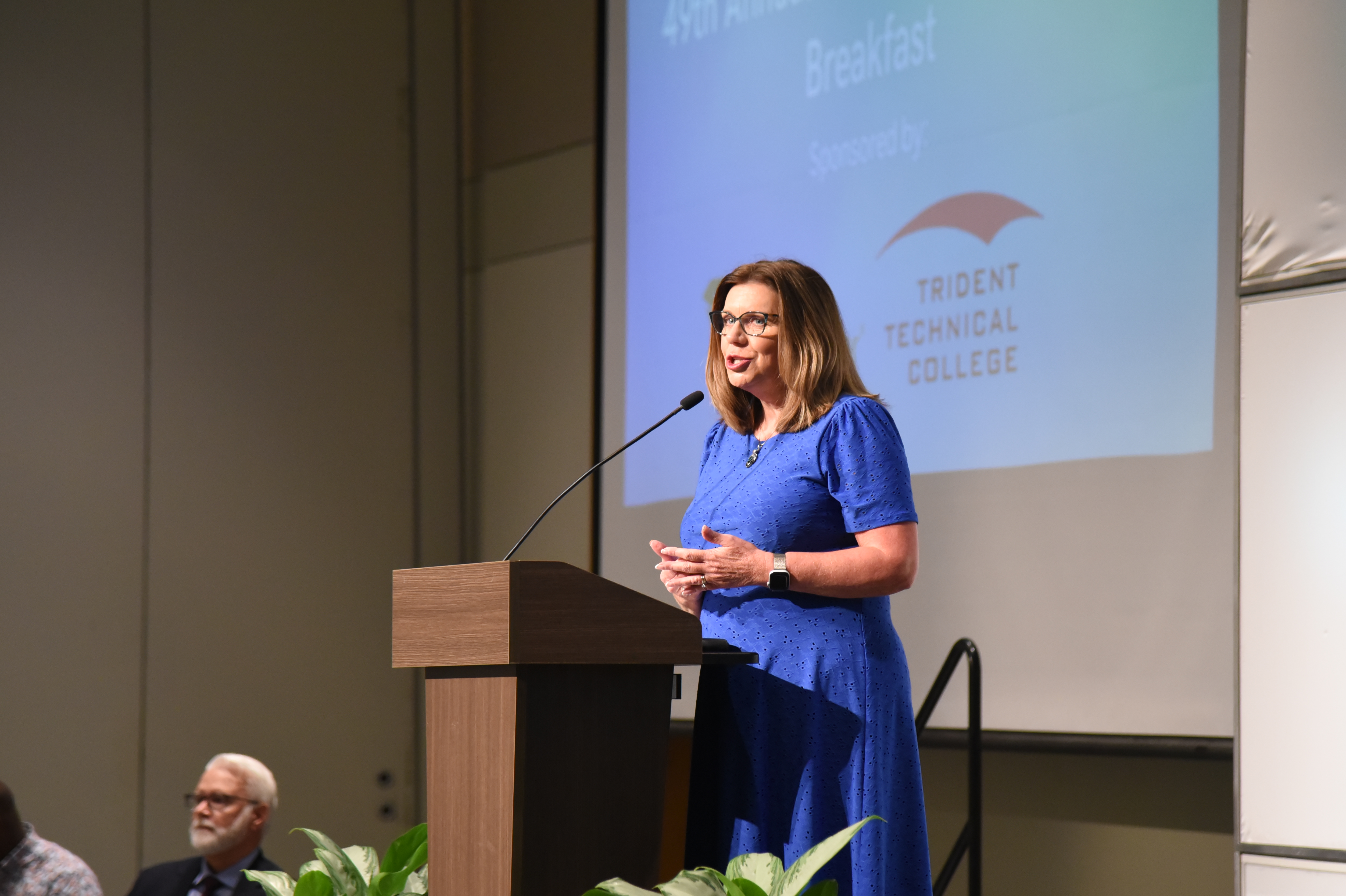 Woman in a blue dress speaking at a podium with a microphone, a large screen behind.