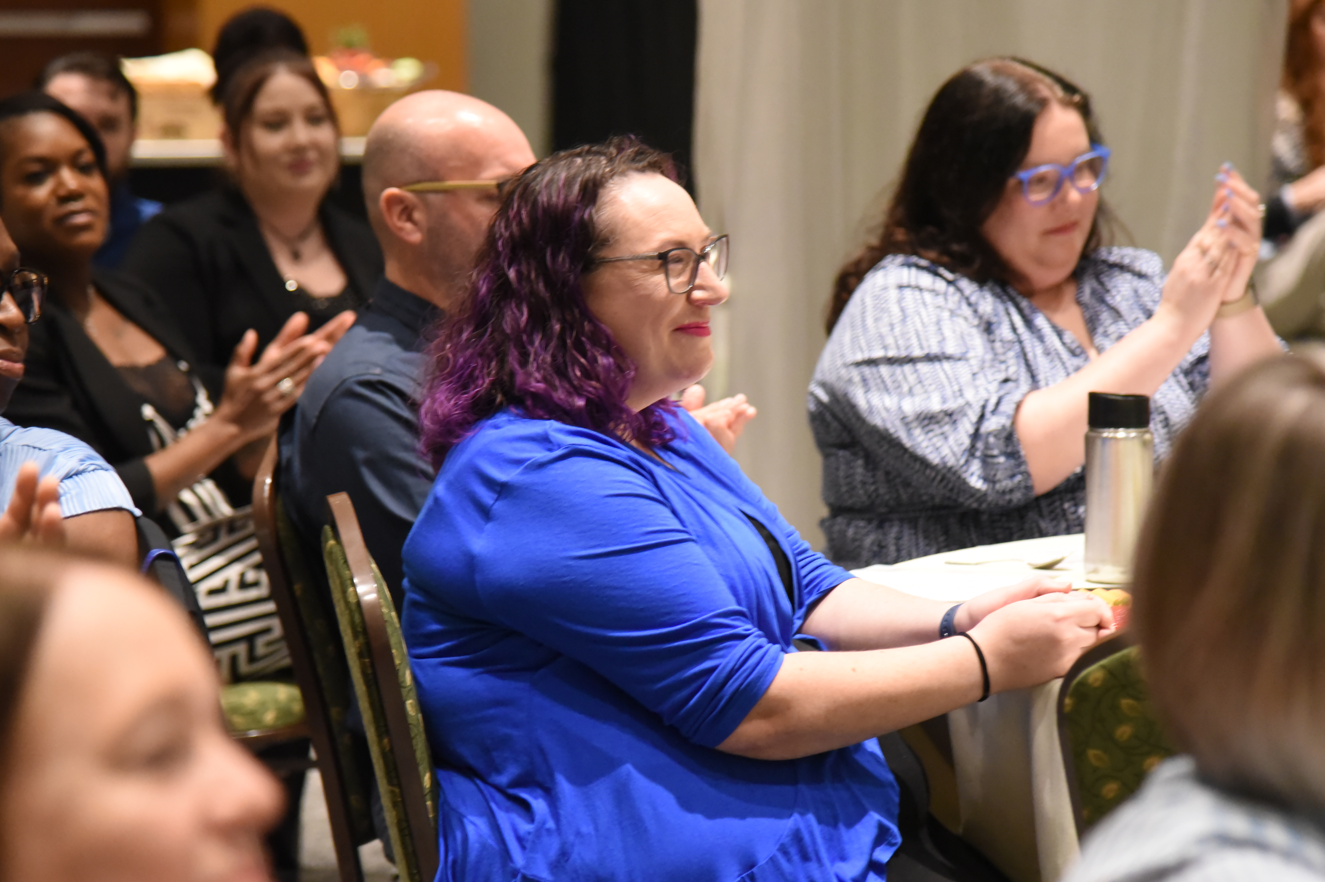 A room filled with people; one woman in blue sits at a table, clapping while others around her do the same.