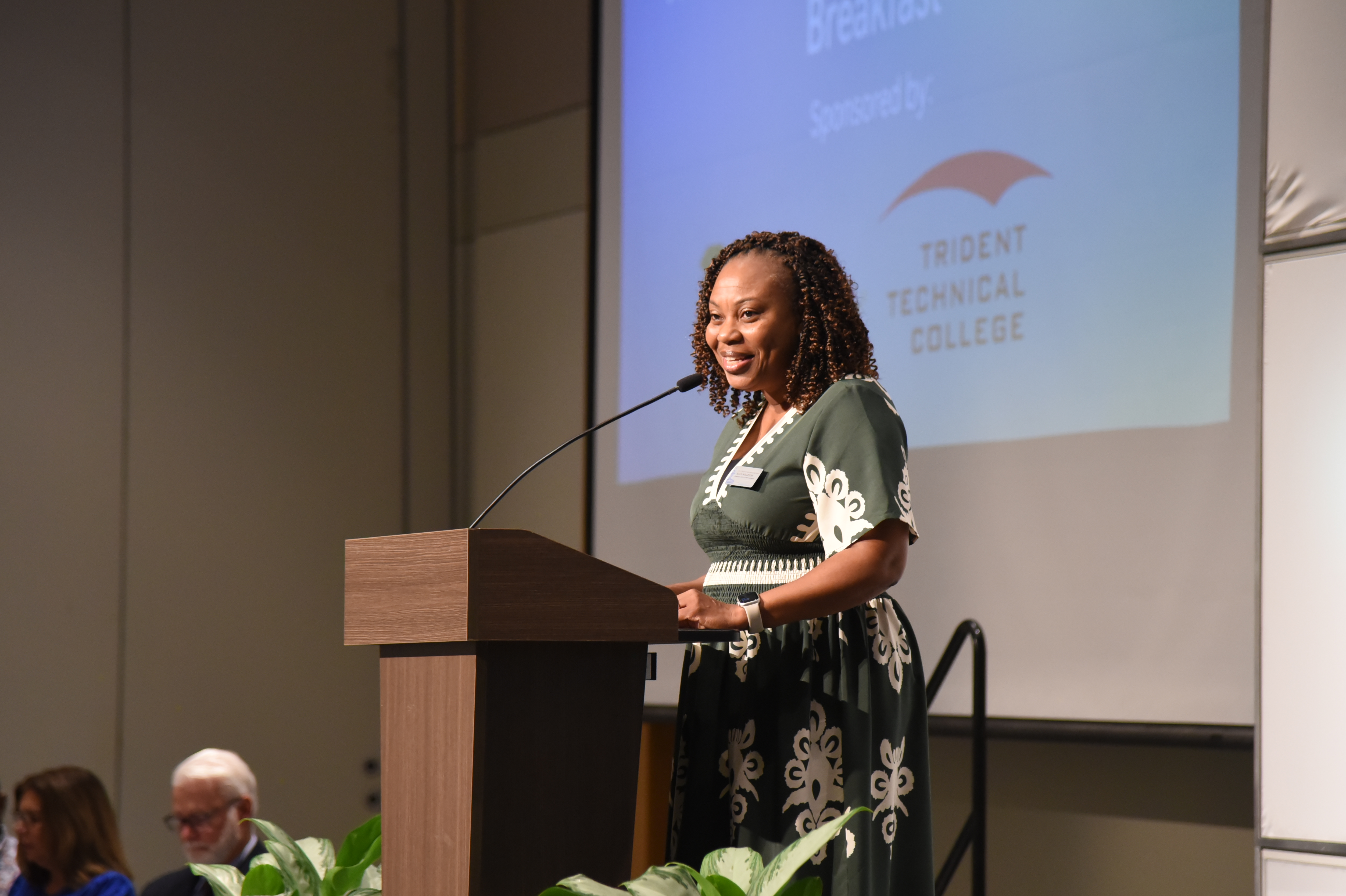 A woman in green speaks at a podium with a microphone. Behind her, a projector screen displays a presentation.