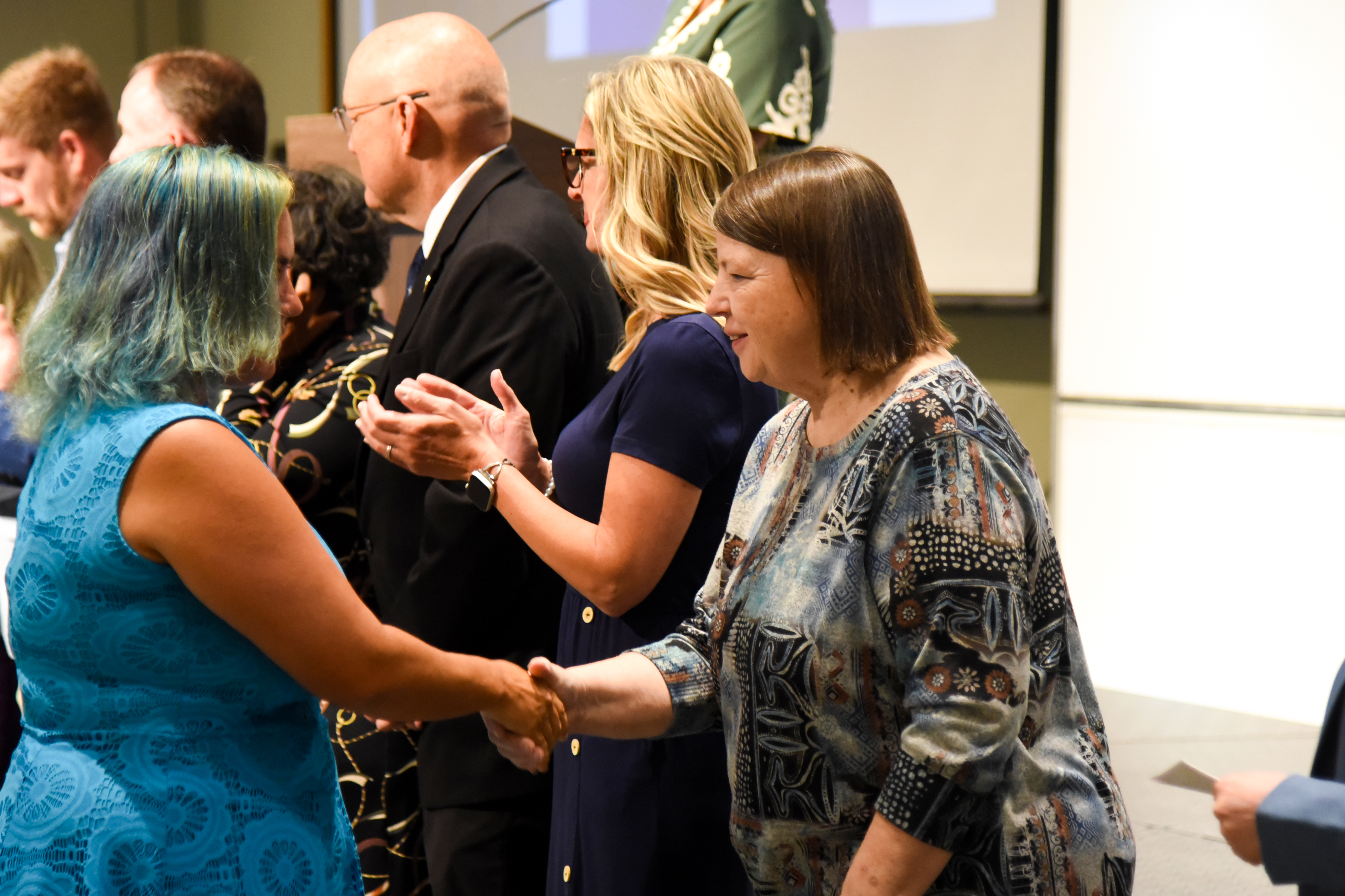 A woman in a blue dress shakes hands with a woman in a patterned shirt at an event.