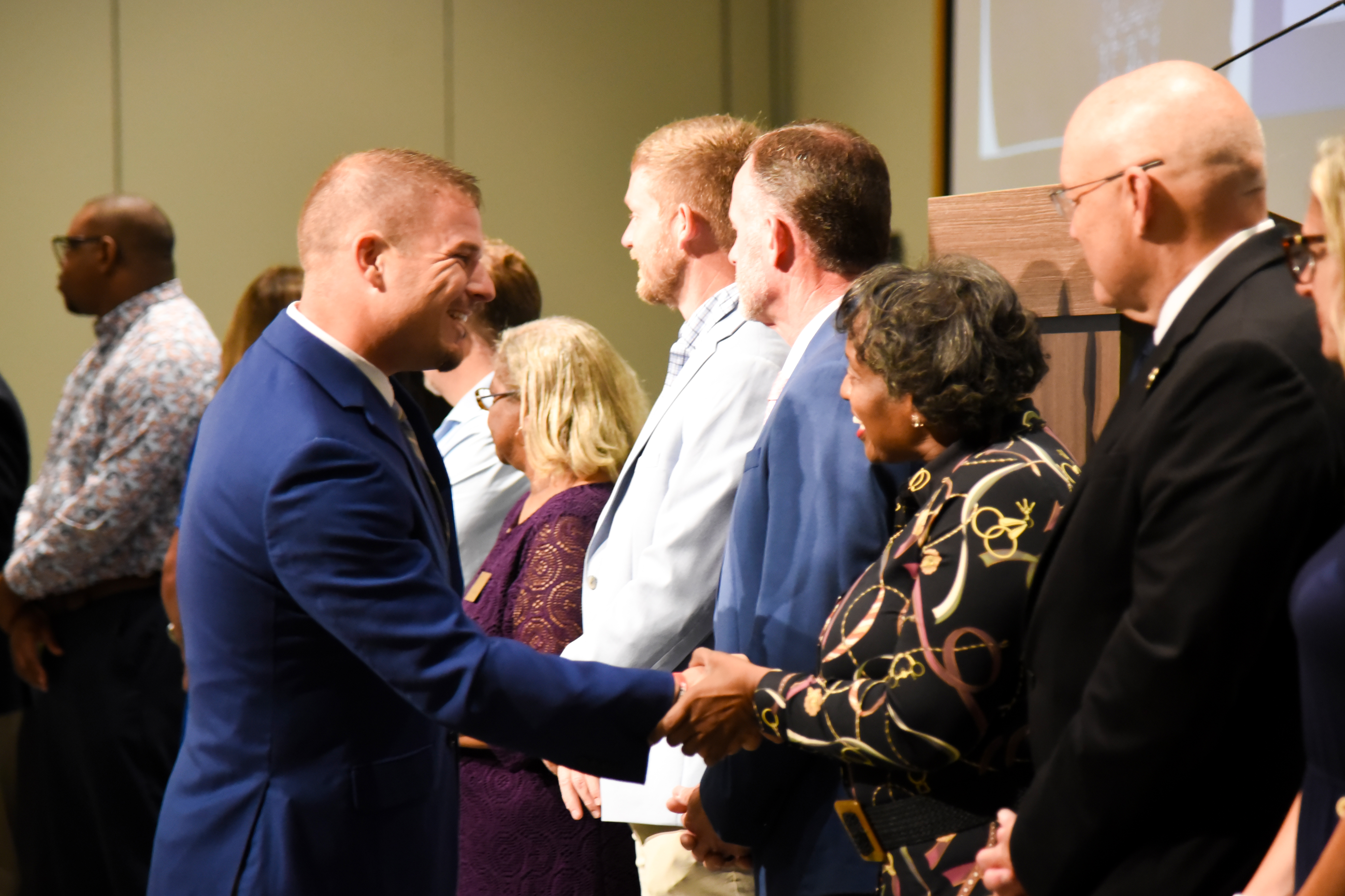 A group of people in formal attire, smiling and shaking hands, with a woman in the center wearing a patterned blouse.