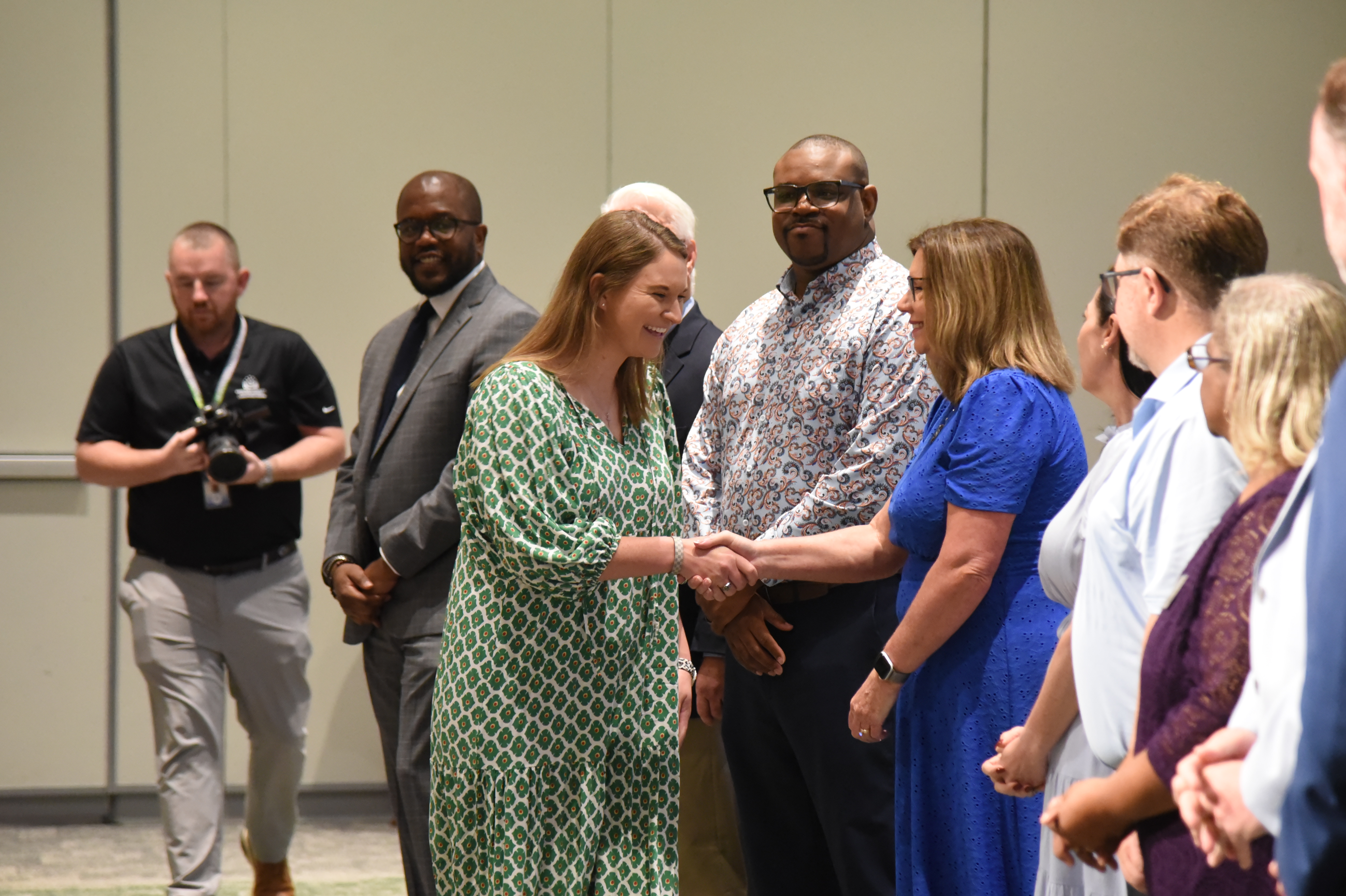 A group of people stand in a room. A woman in a green dress shakes hands with a man in a blue dress.