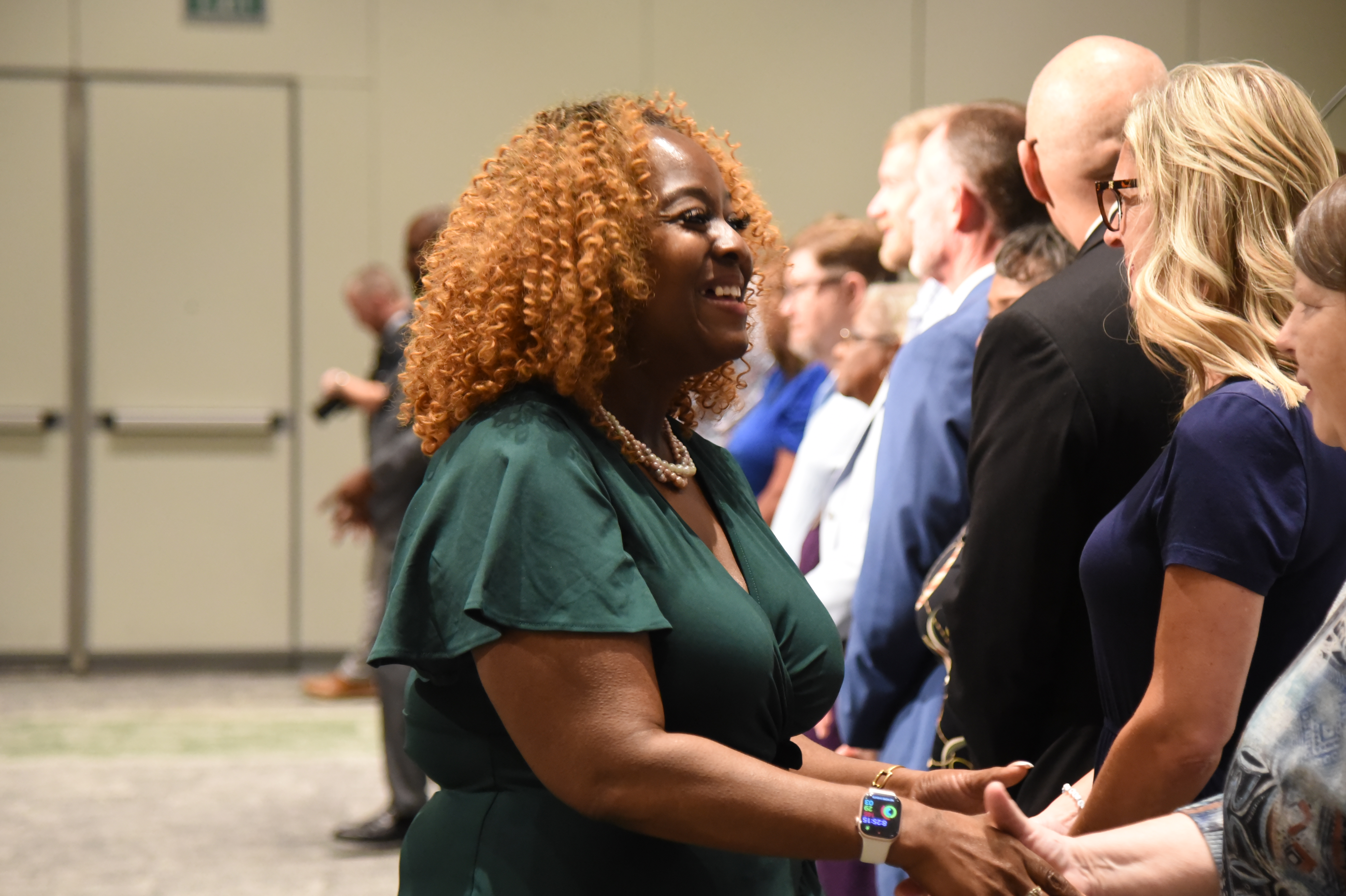 A woman in a green dress stands smiling in a crowd of people in a conference hall.
