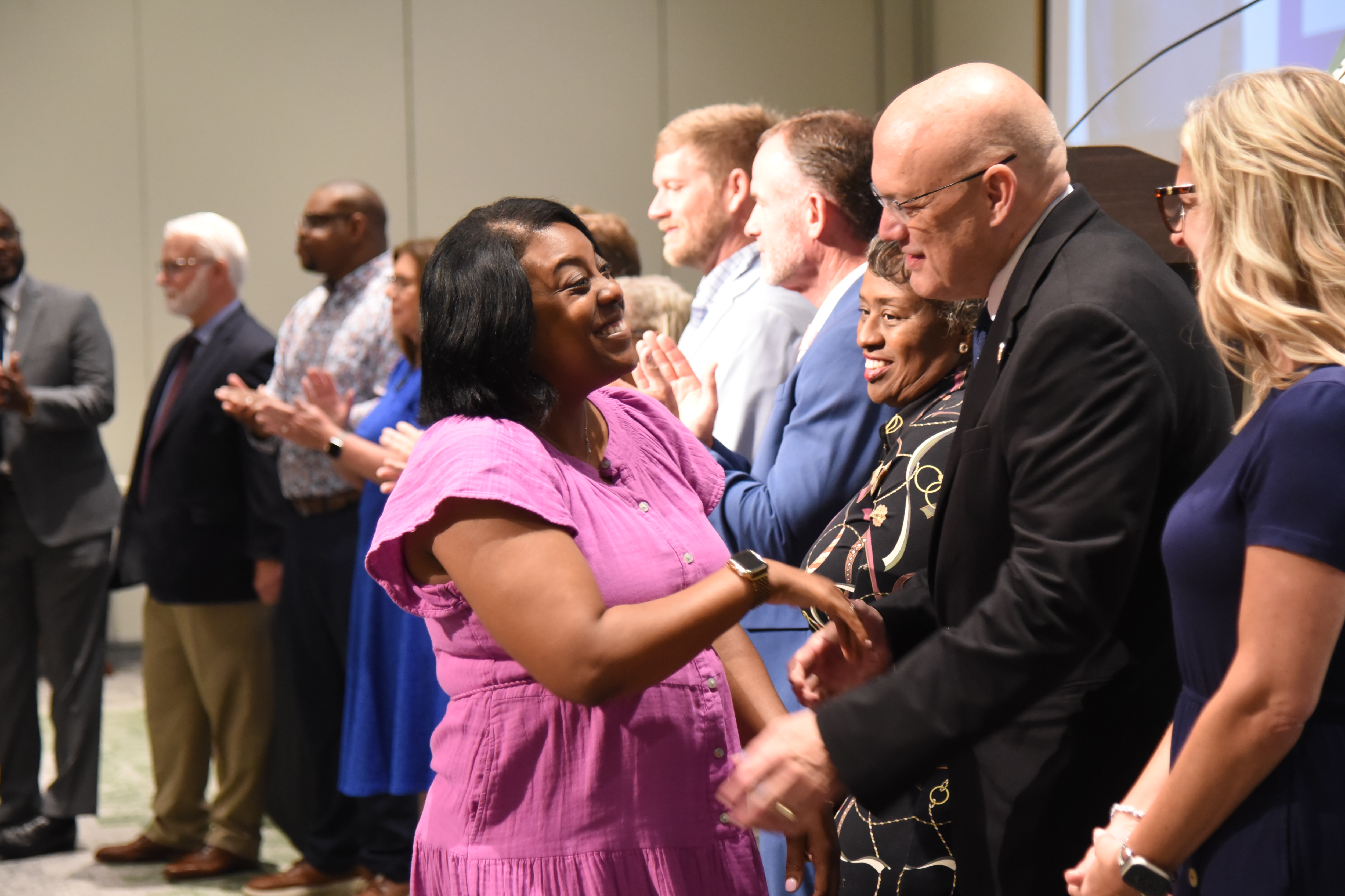 A group of people stand in a line, clapping. A woman in a pink dress holds hands with a man in a black suit.