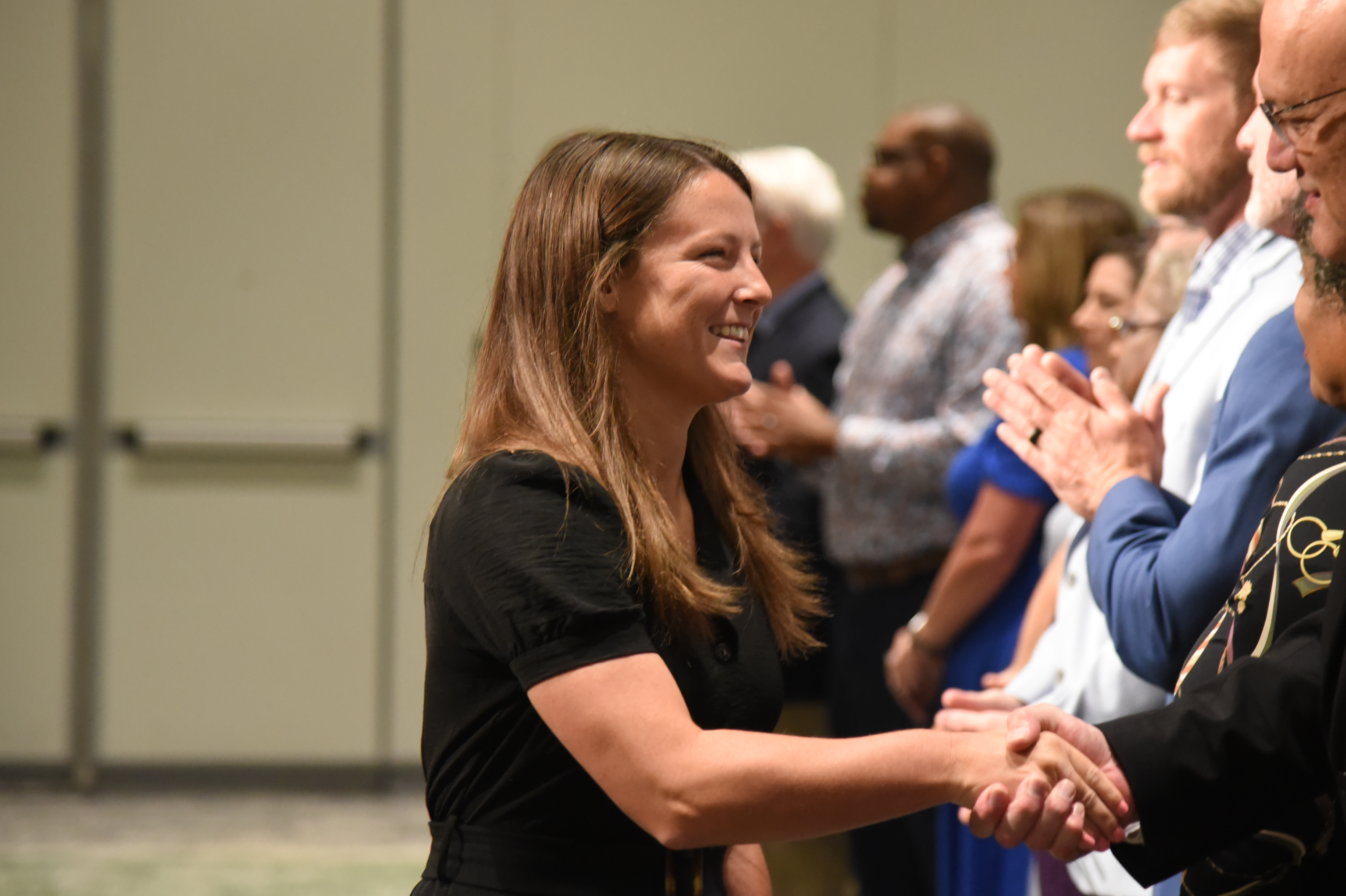 A smiling woman in a black shirt shakes hands with a man while others in the background clap.
