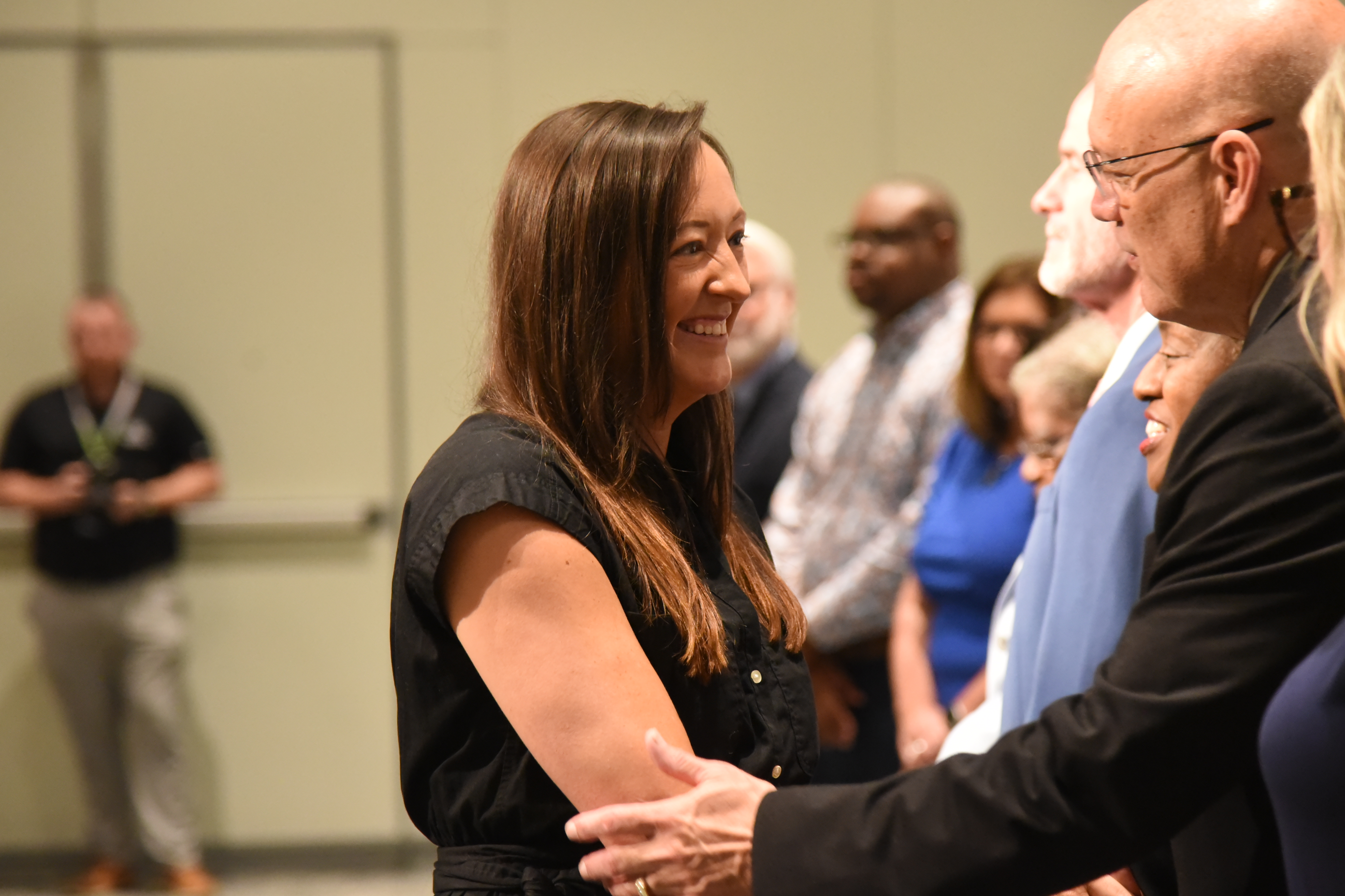 People in a room, a woman and man smiling and shaking hands, others standing behind them.