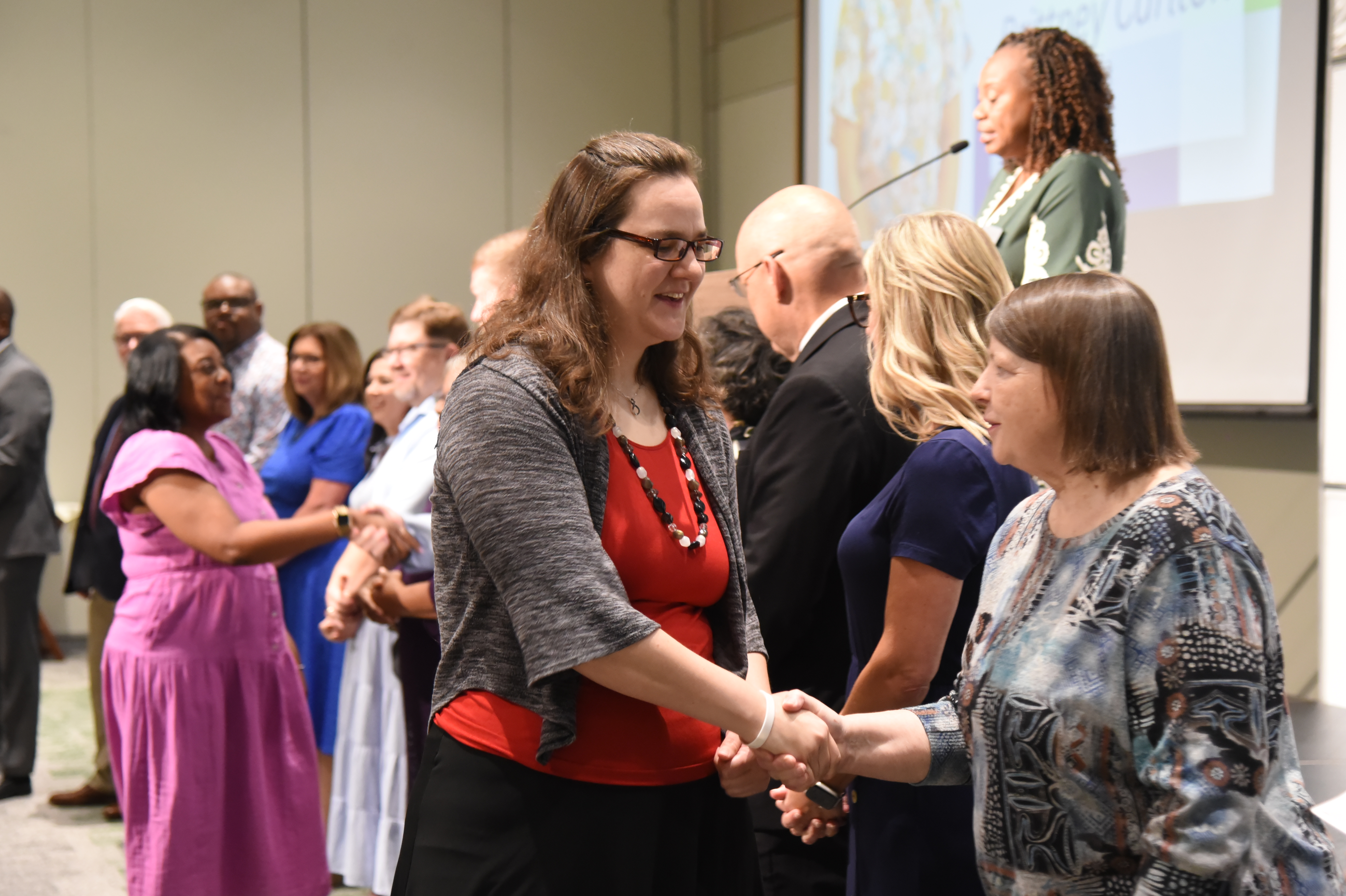 People in formal attire, some in dresses, gather in a room, shaking hands in front of a projector screen.