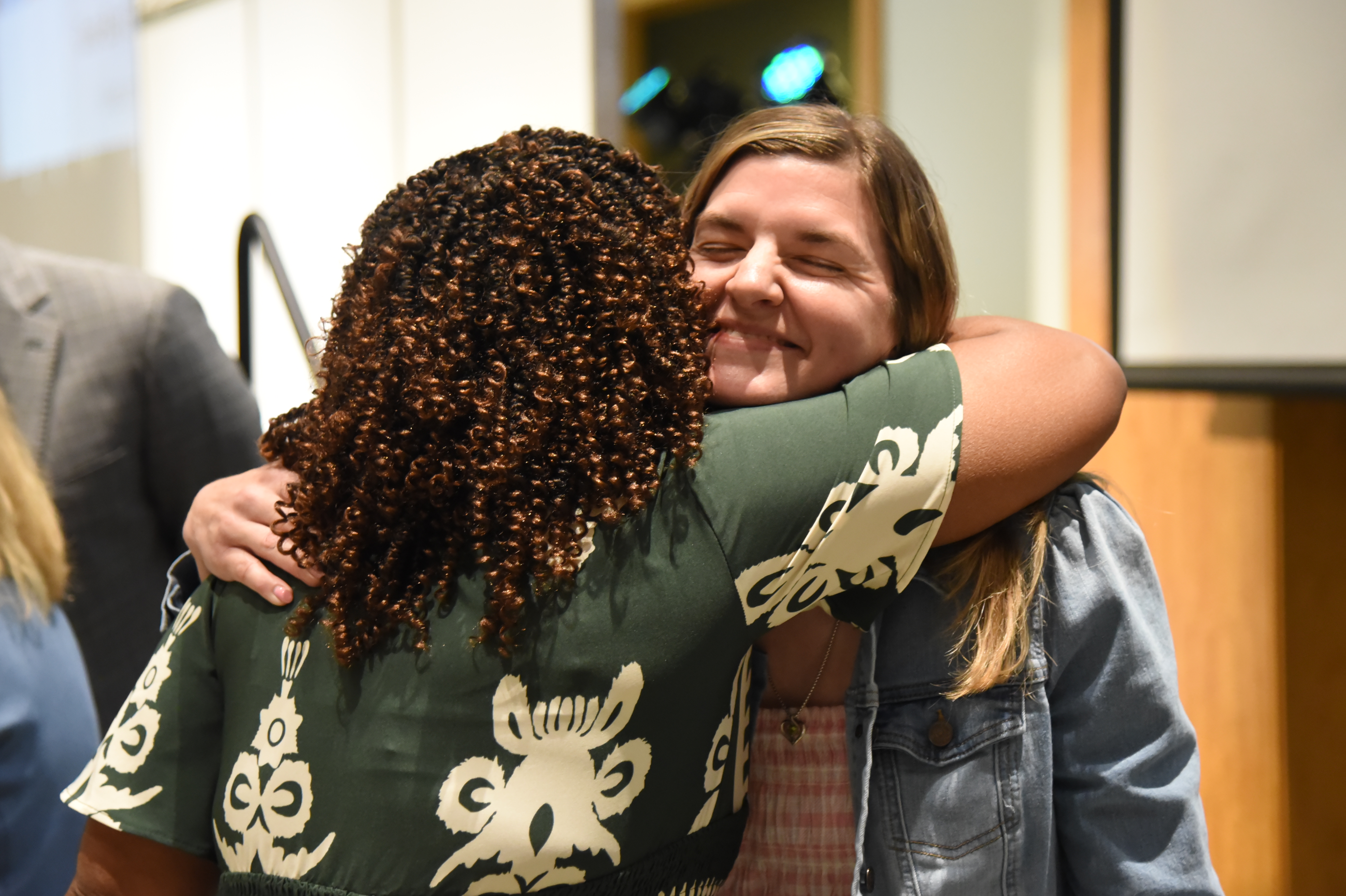 Two women embrace in a hug. One has curly hair, wearing a green shirt. The other has straight hair, wearing a denim jacket.