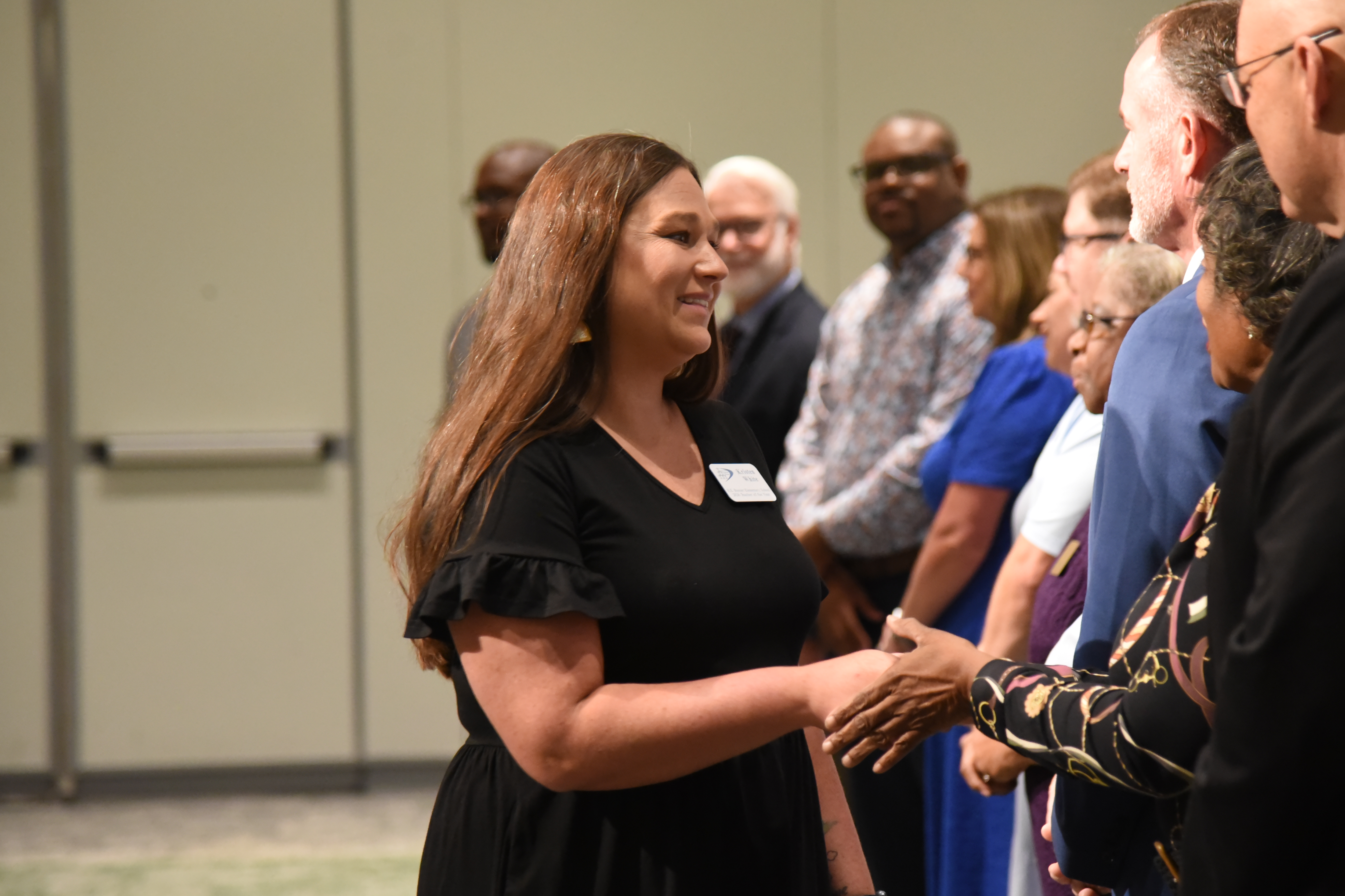 A woman in a black dress greets someone with a handshake, surrounded by other people at an indoor event.