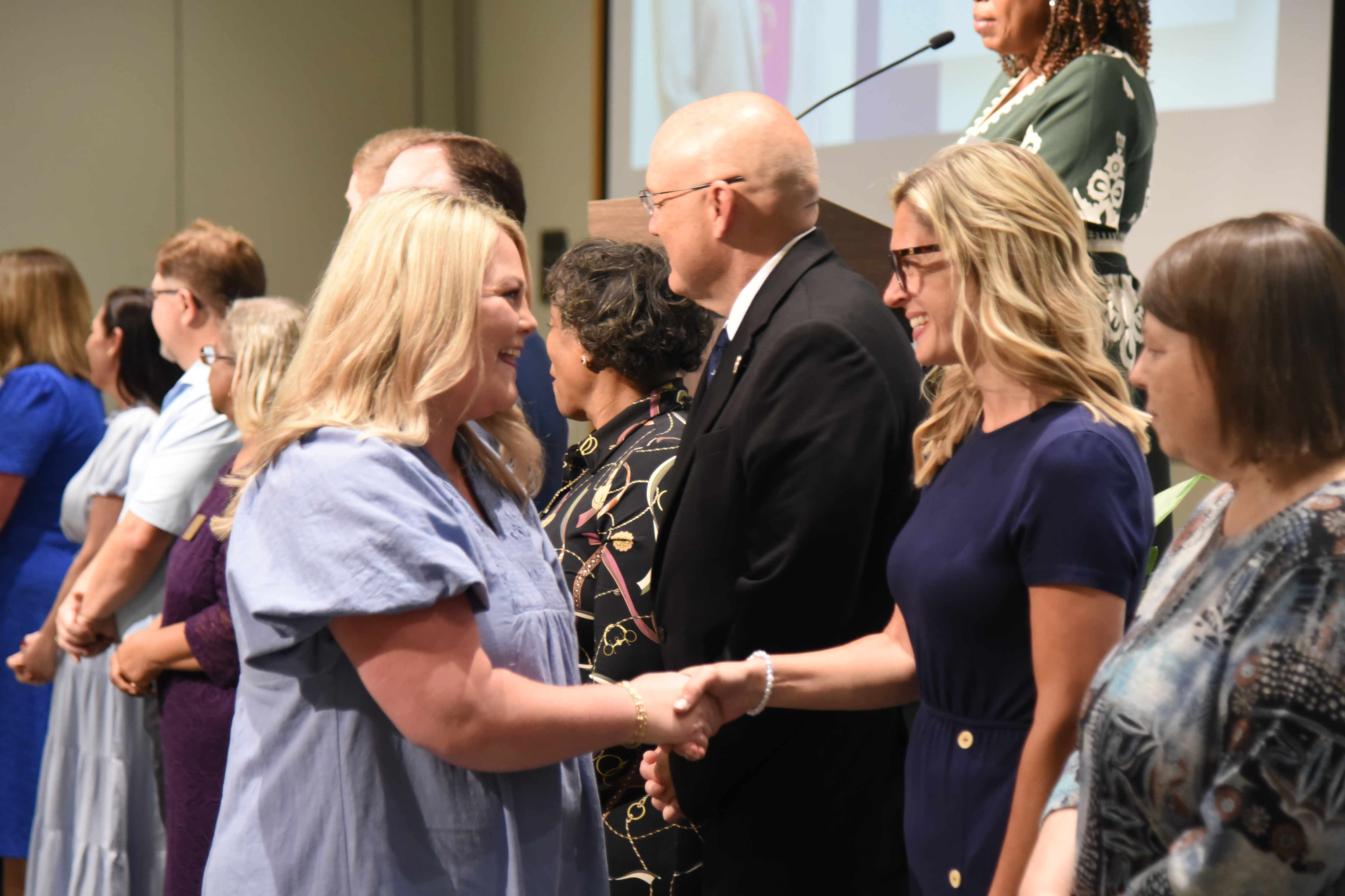 A group of people, including men and women, dressed formally, stand together. Some shake hands.