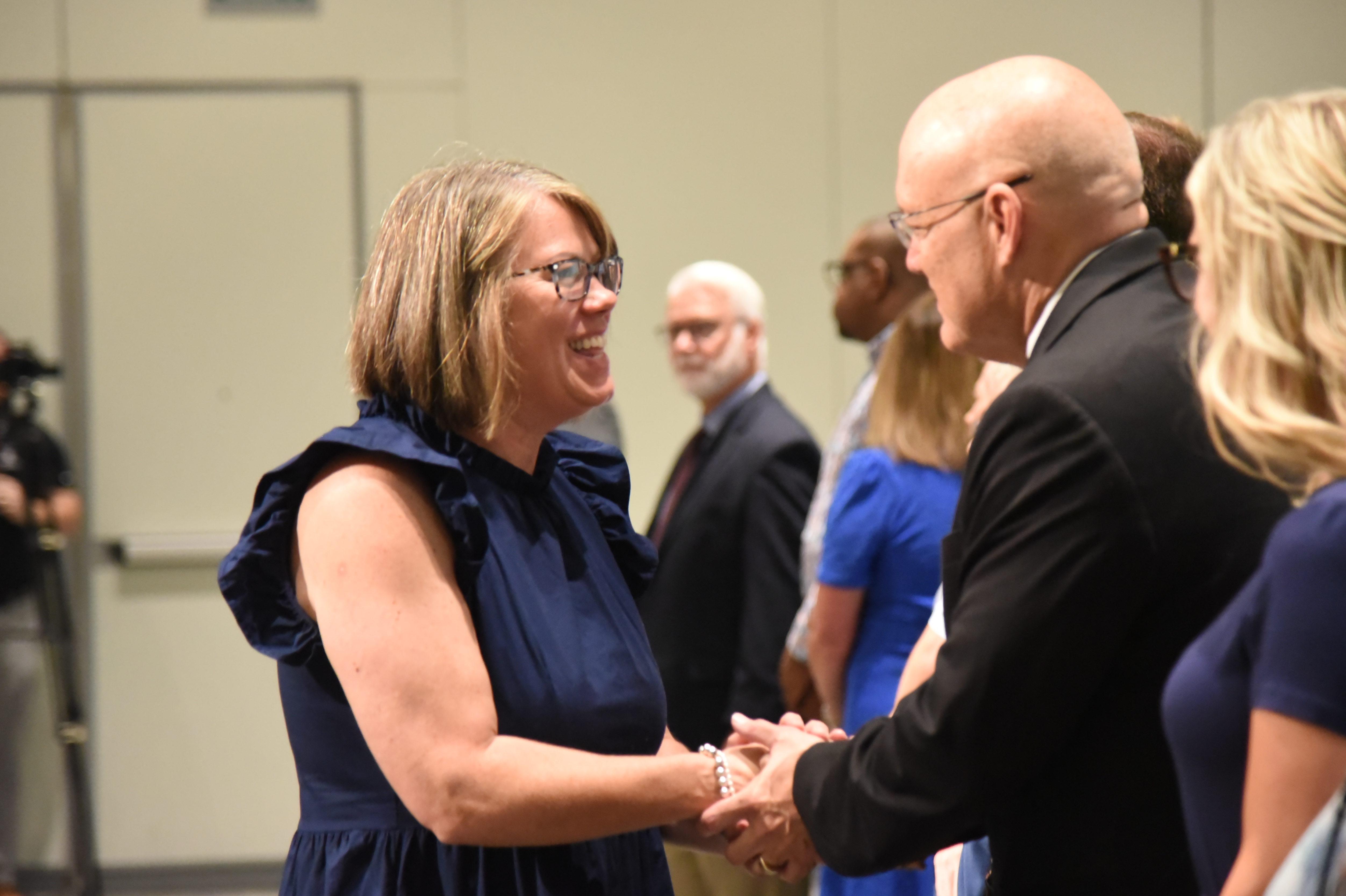 People shake hands, smile, and converse in an indoor event; a woman in a blue dress and man in a black suit.