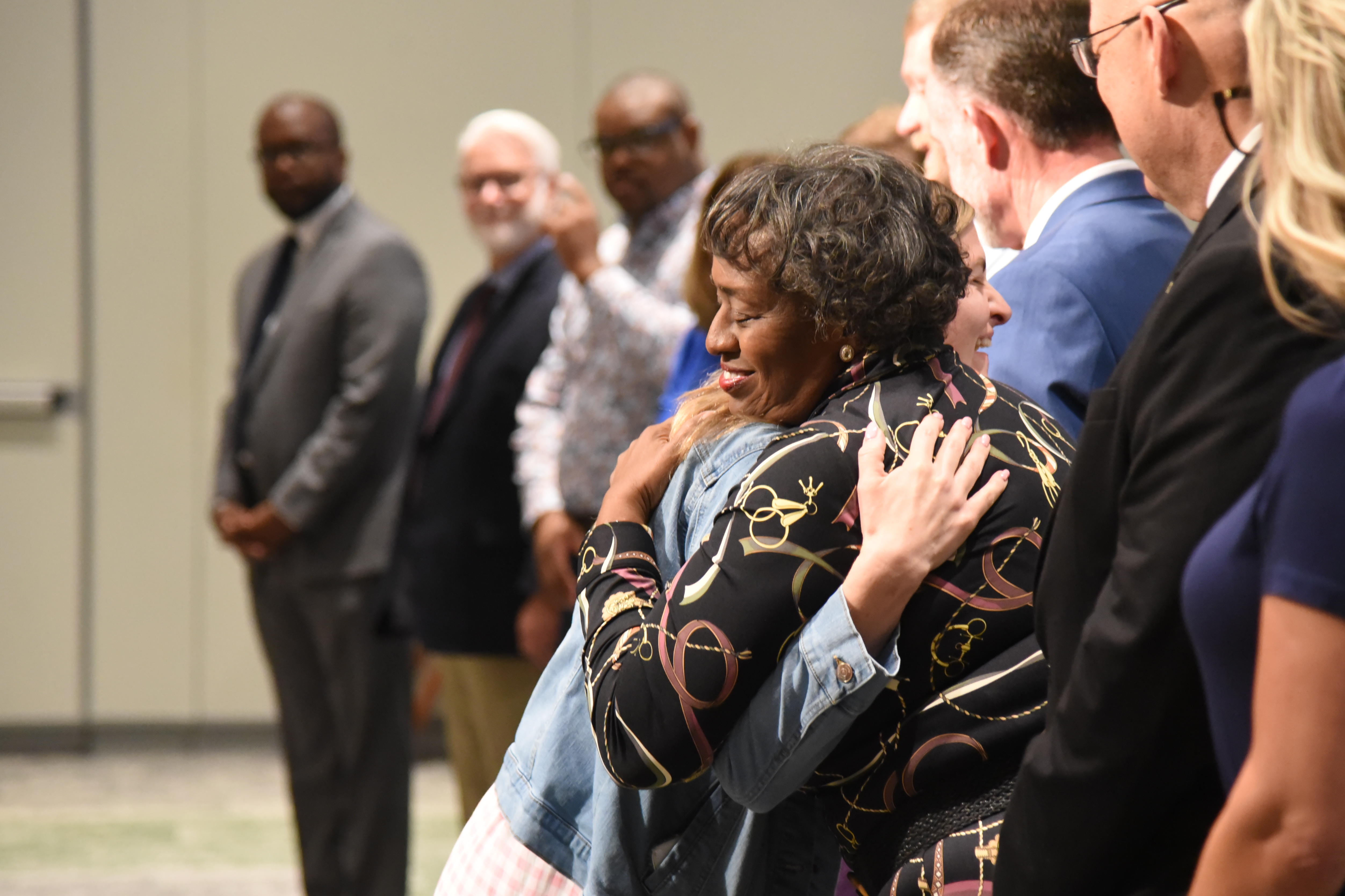 People stand and gather in a room. A woman in a floral top embraces another person.