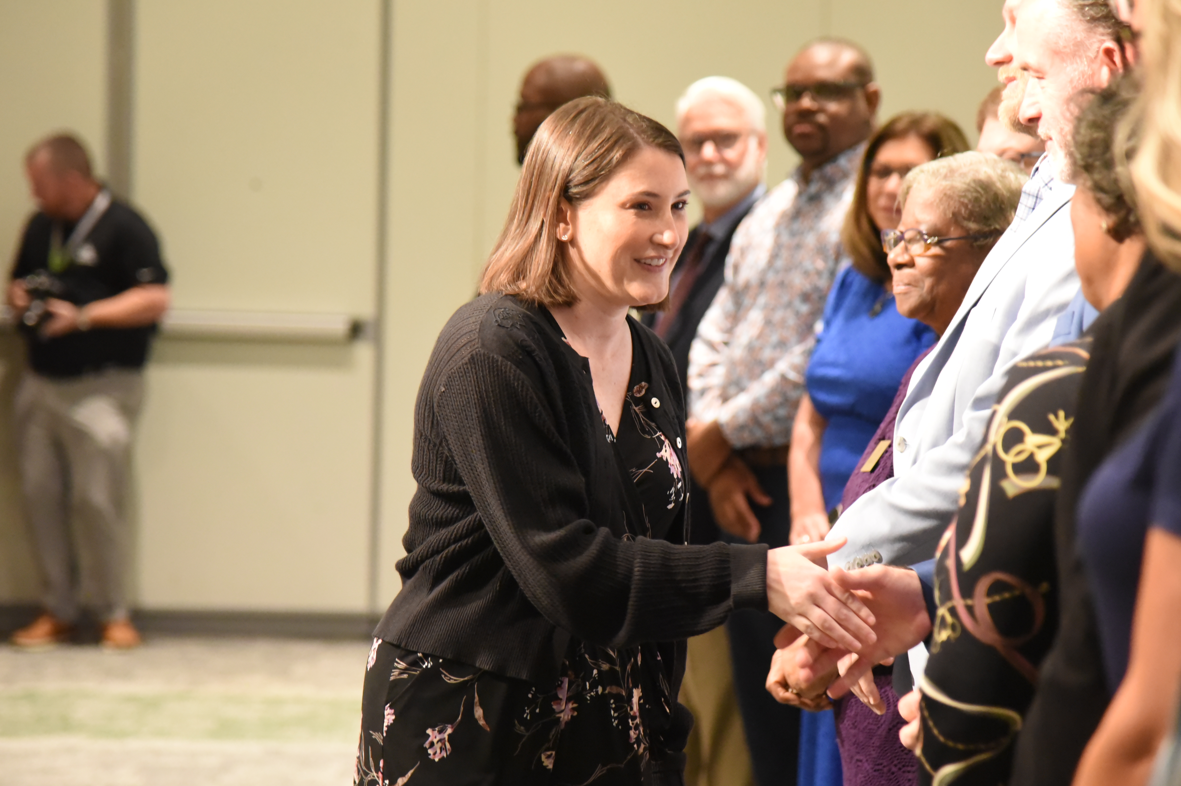 A woman shakes hands with a group of adults, smiling. A man stands behind her, and others look on.