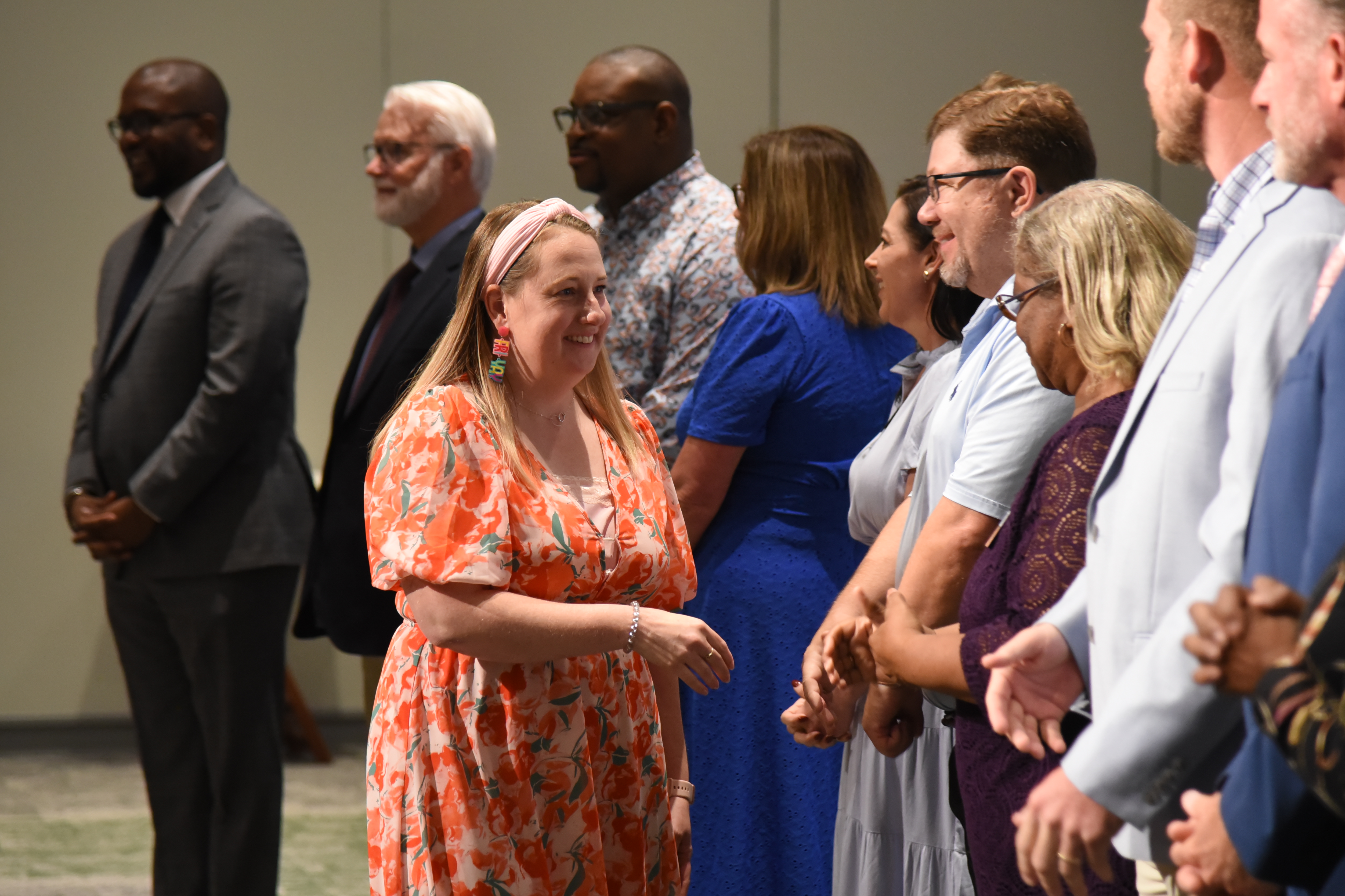 A group of people stands in a room, some wearing formal attire. A woman in an orange dress smiles.