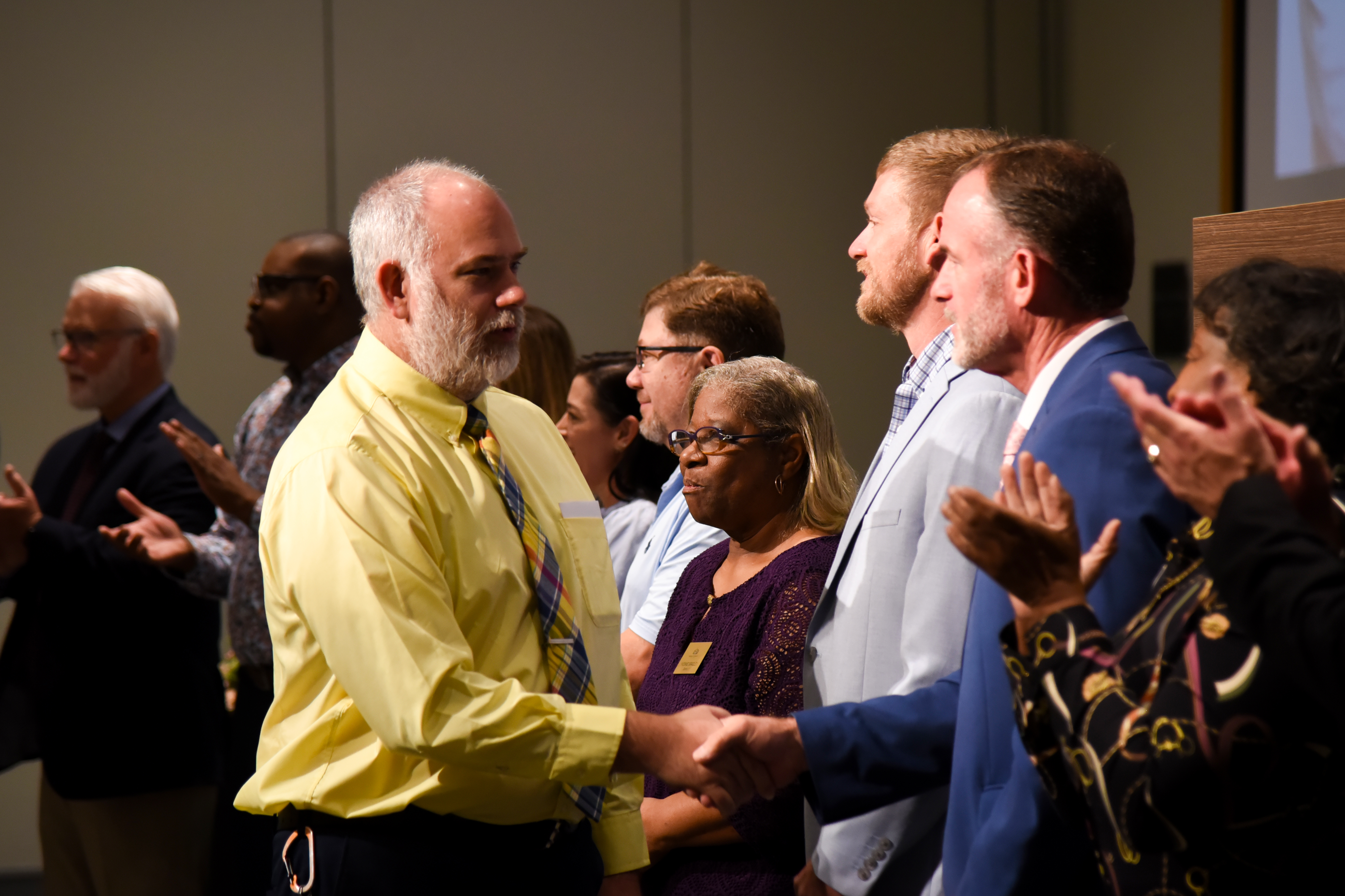 A group of people in a room. A man in a yellow shirt shakes hands with another man.