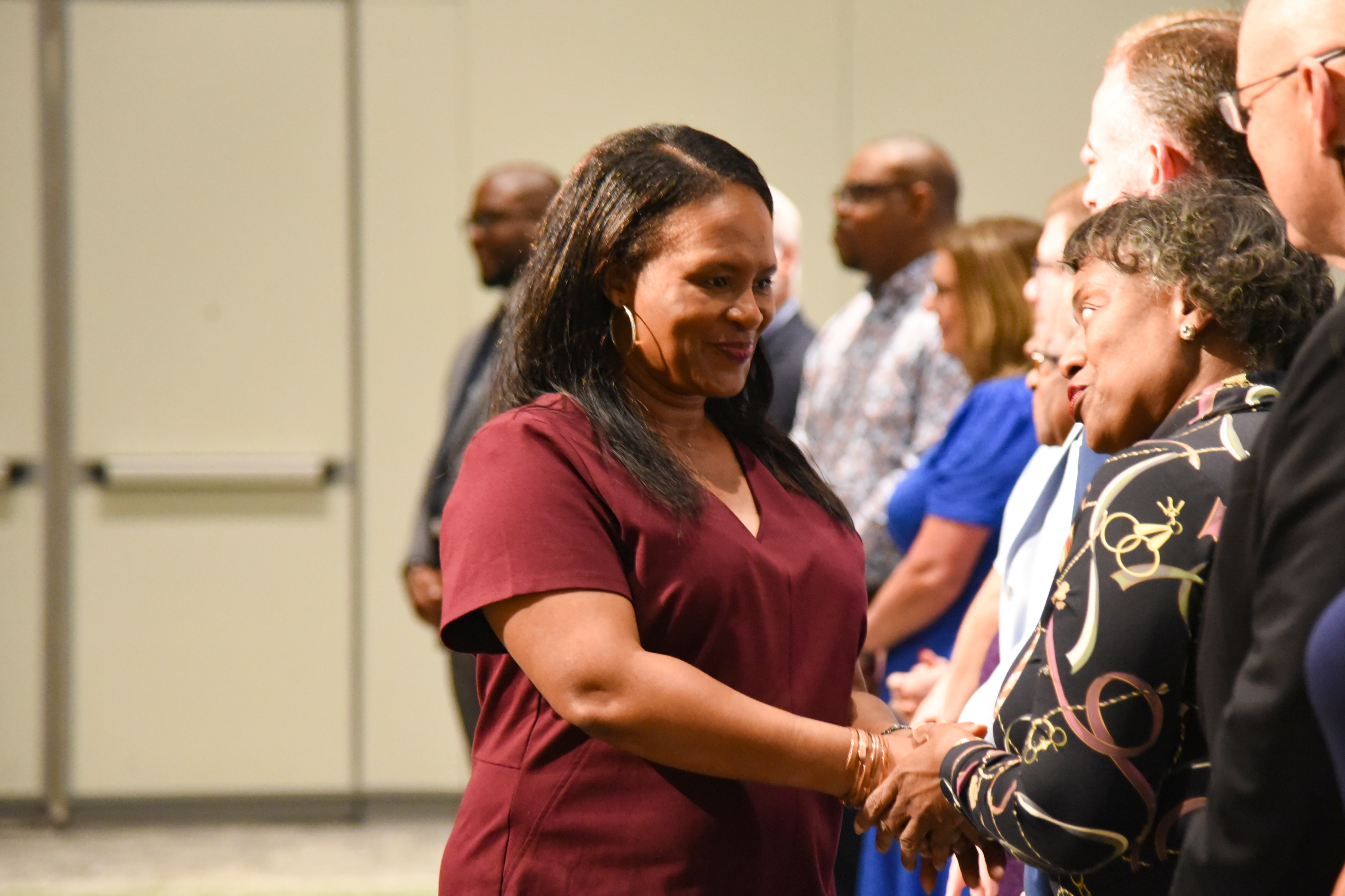 People stand in a room with some smiling. A woman in a maroon shirt shakes hands with a woman in a patterned shirt.