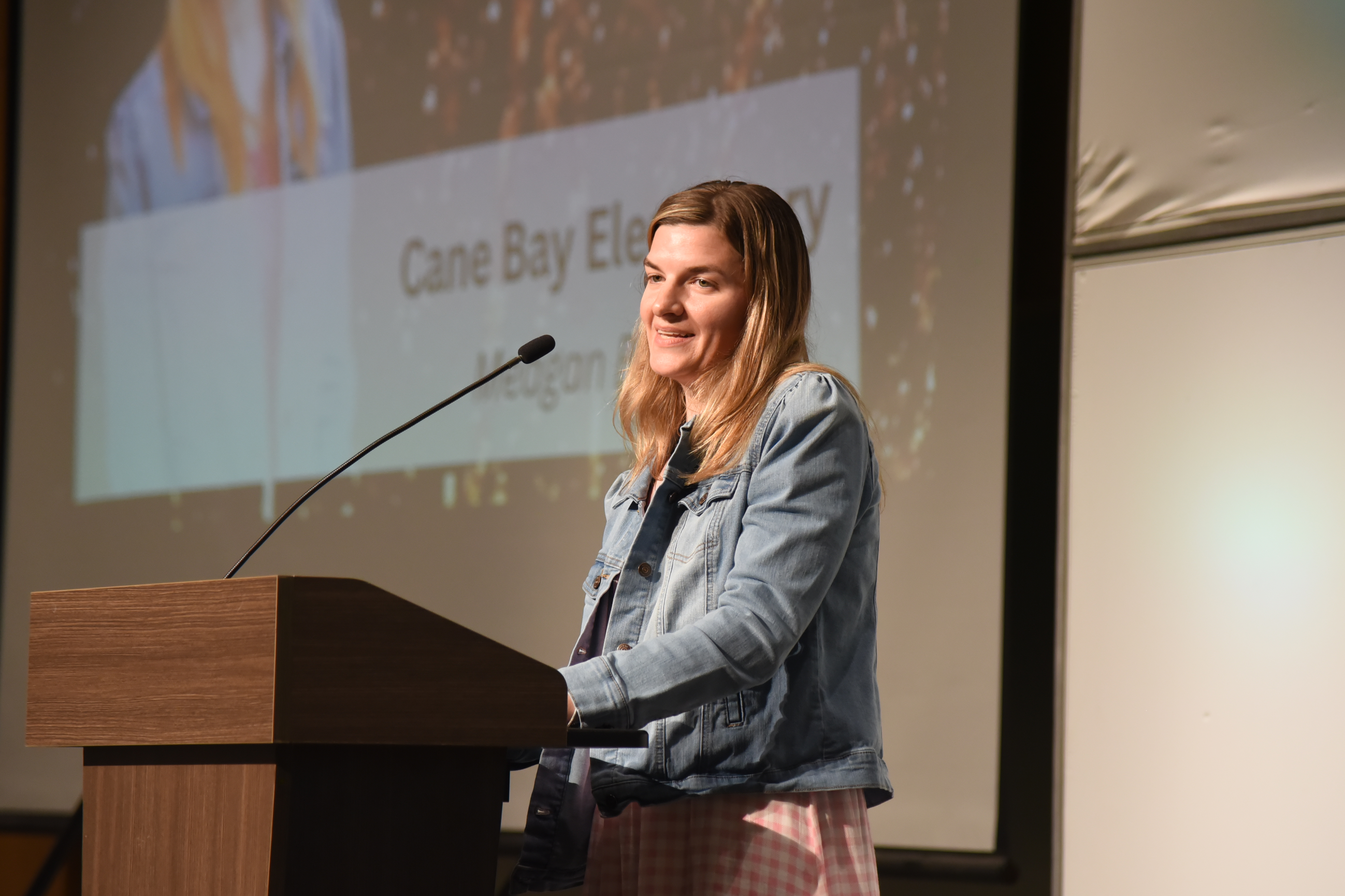 A woman speaks at a podium in a conference room. The projector screen reads "Cane Bay Elementary."