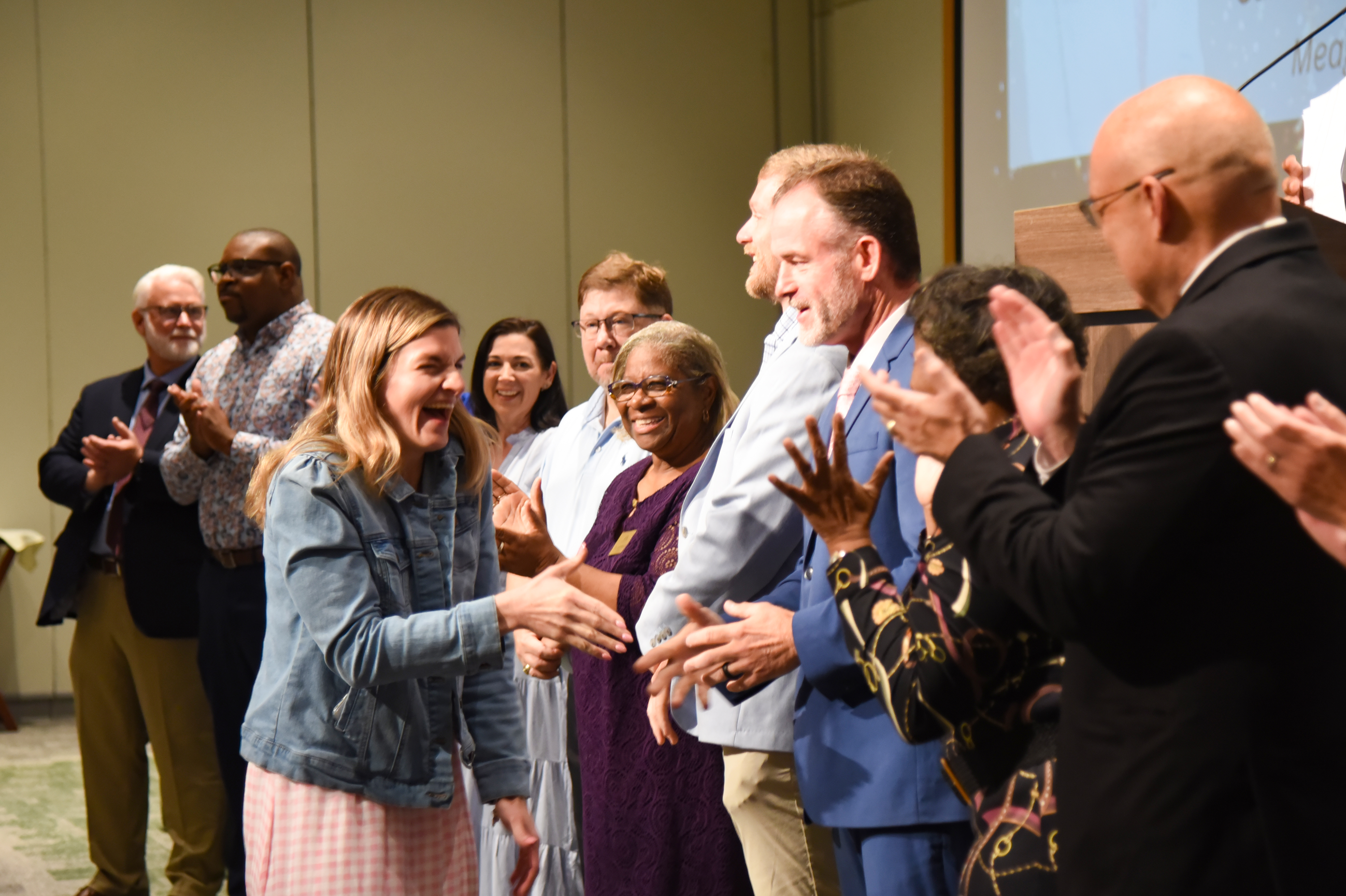 A group of adults in formal and casual attire clapping in an indoor setting with a projector screen in the background.